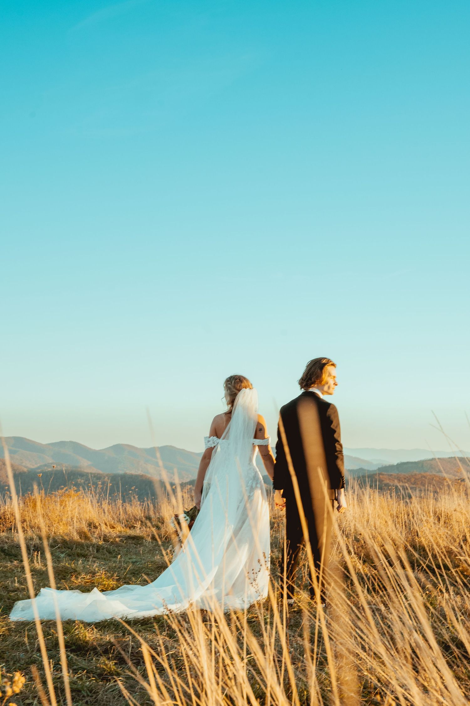 Bride and groom standing in golden mountain field with flowing white dress and veil.