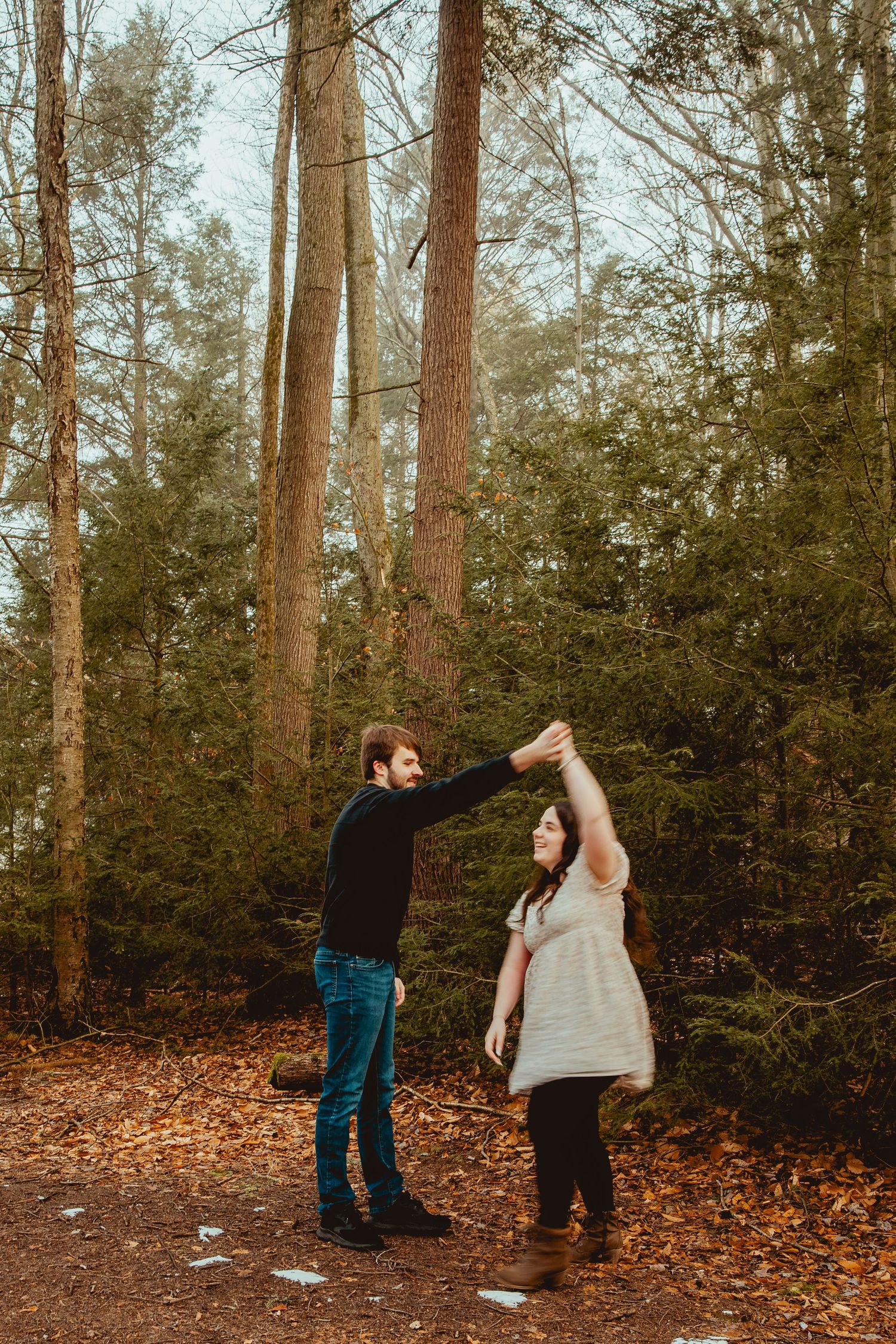 Two people playfully interact in a wooded area with fallen leaves and evergreen trees in the background.
