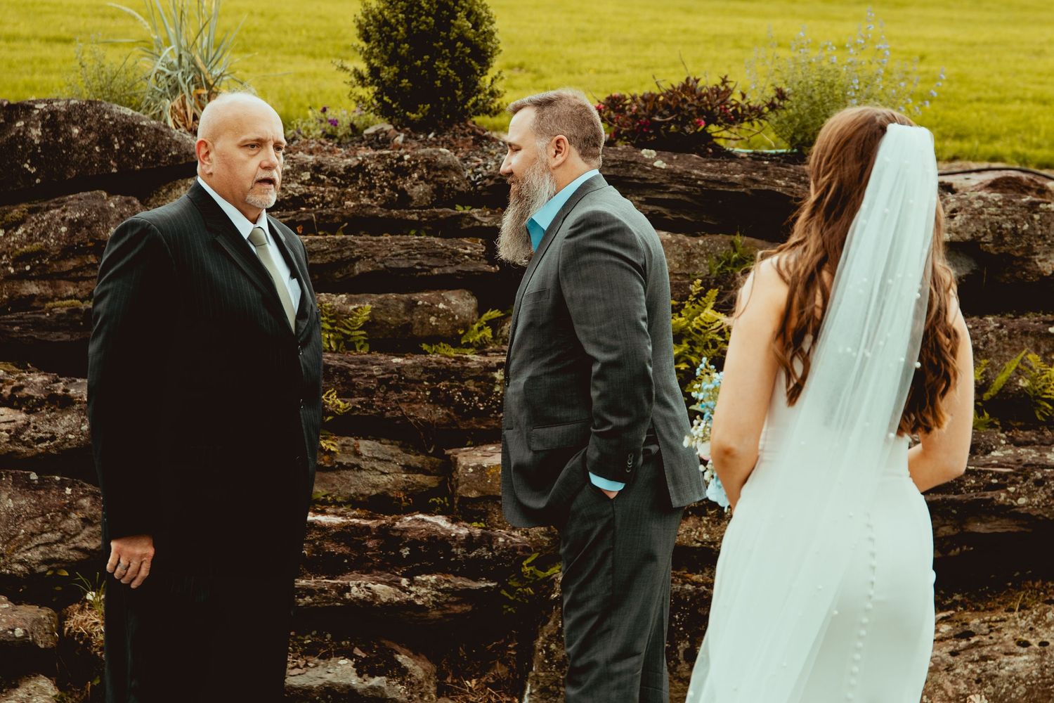 Wedding party members interacting and laughing together near a rustic stone wall outdoors.