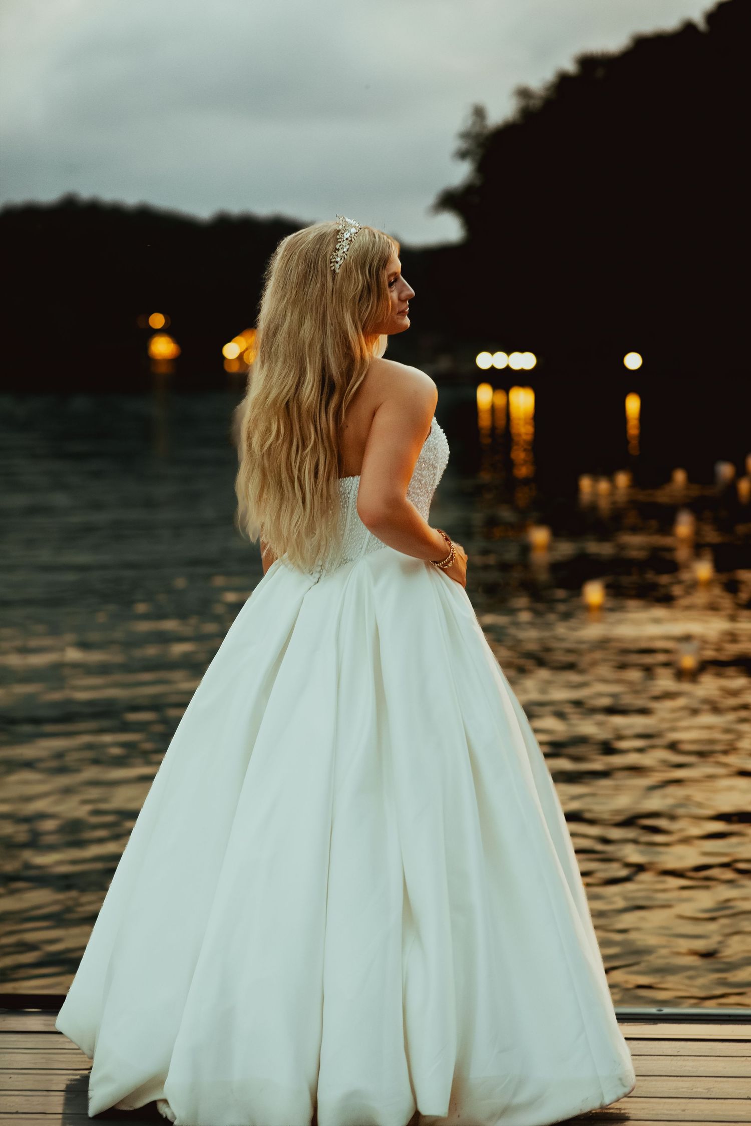 Bride in a stunning white ball gown stands on a dock at dusk with bokeh lights reflecting on water.