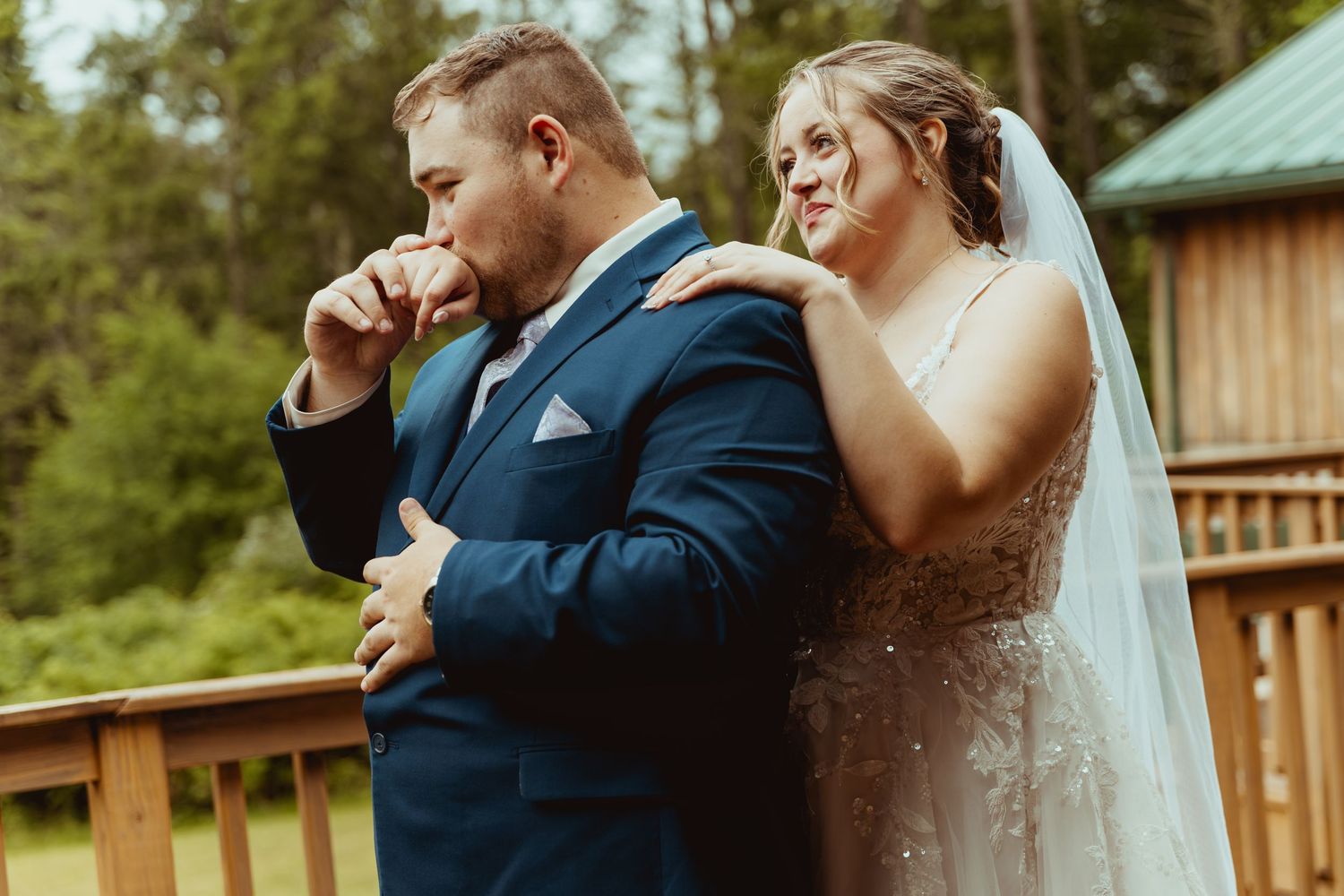 Bride and groom share an emotional first look moment outdoors, with the groom wiping tears as the bride stands behind him.