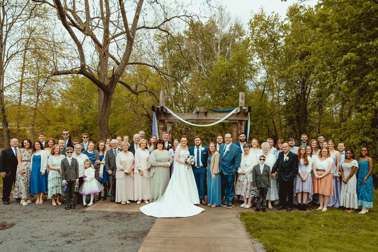 Large wedding party group photo outdoors with bride and groom surrounded by guests near decorated arch.