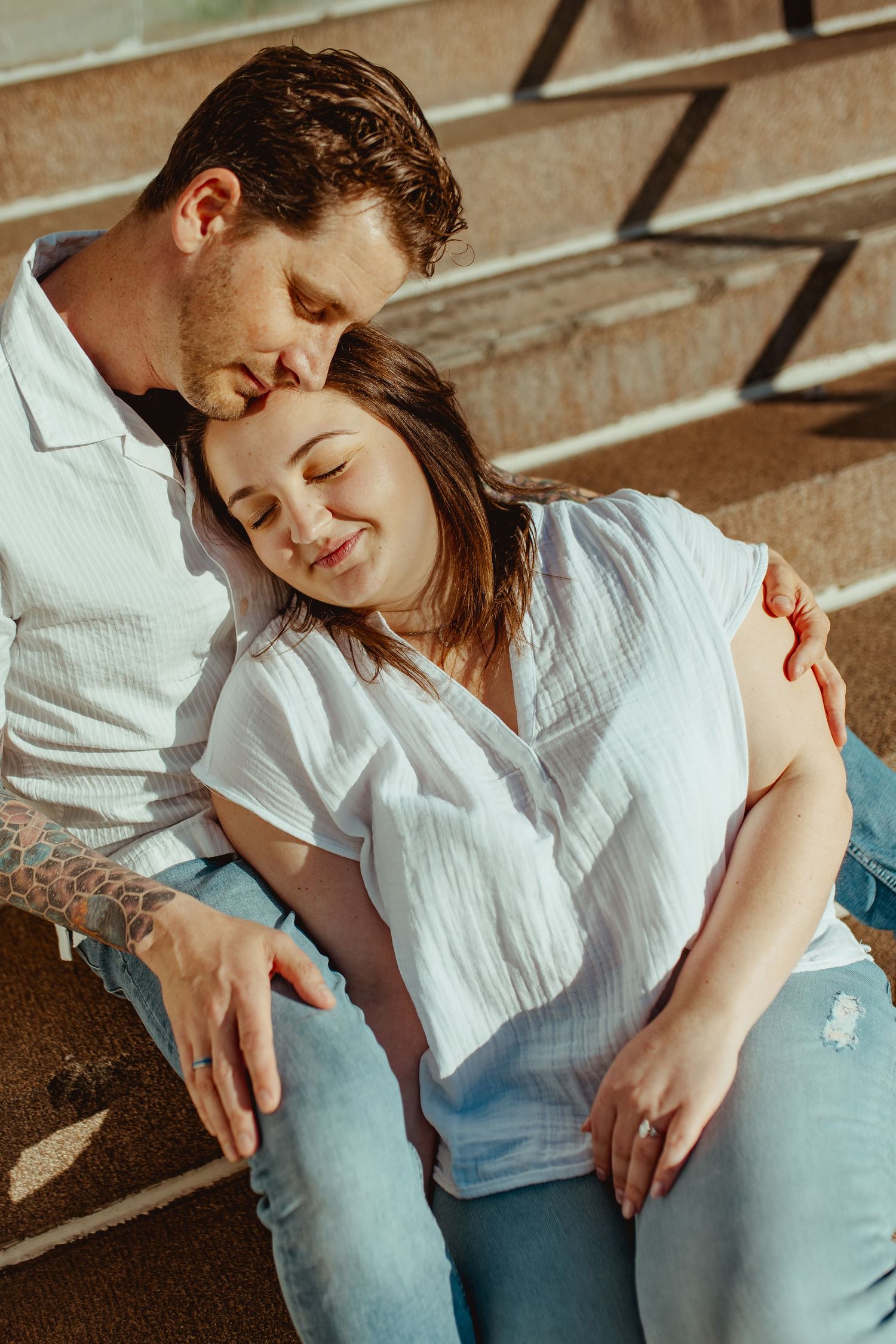 A couple in white shirts and jeans sit on steps, sharing a tender embrace in warm sunlight.