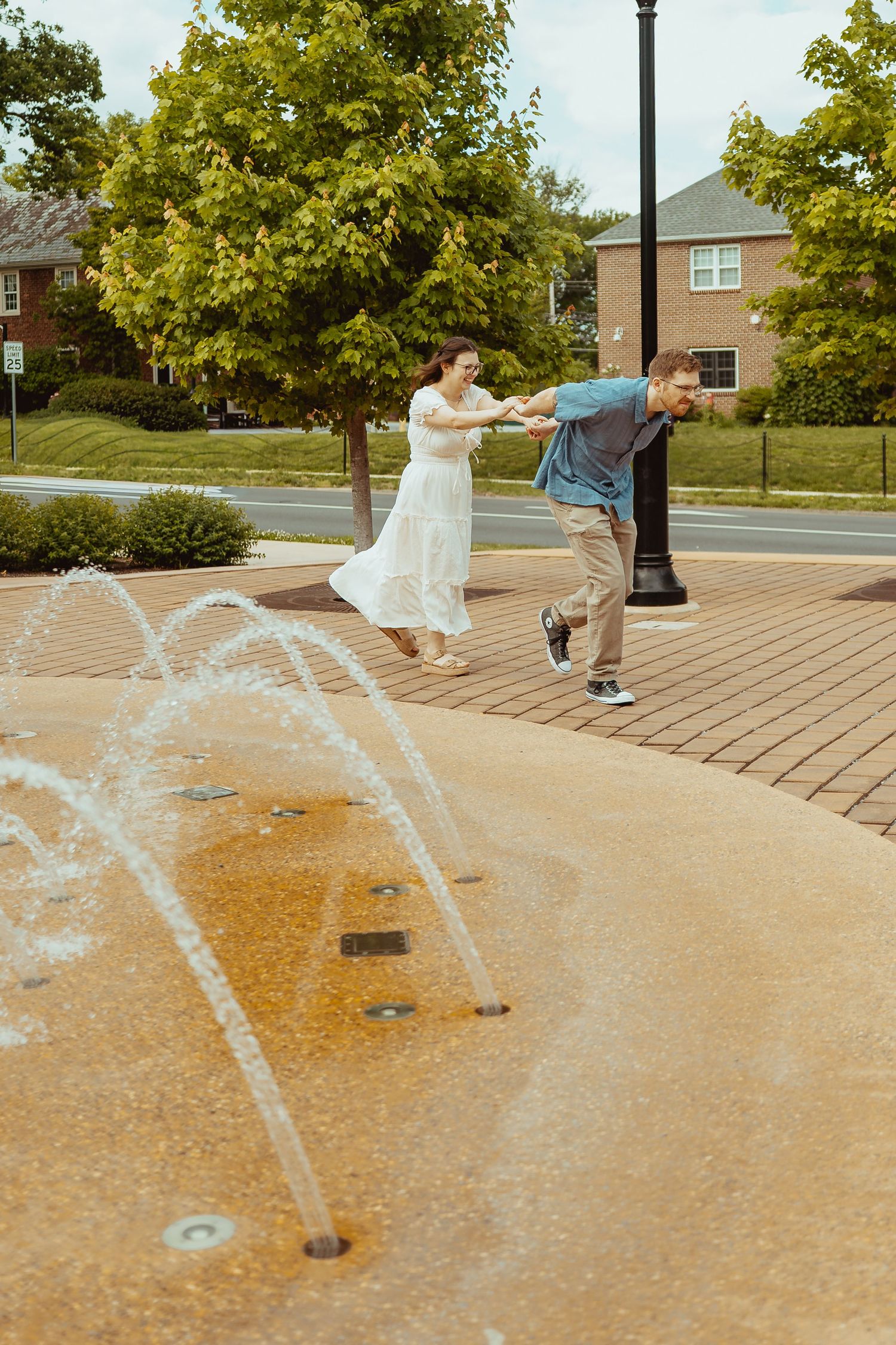 A joyful couple plays near a splash pad fountain, the woman in a white dress holding hands with a man in a blue shirt.