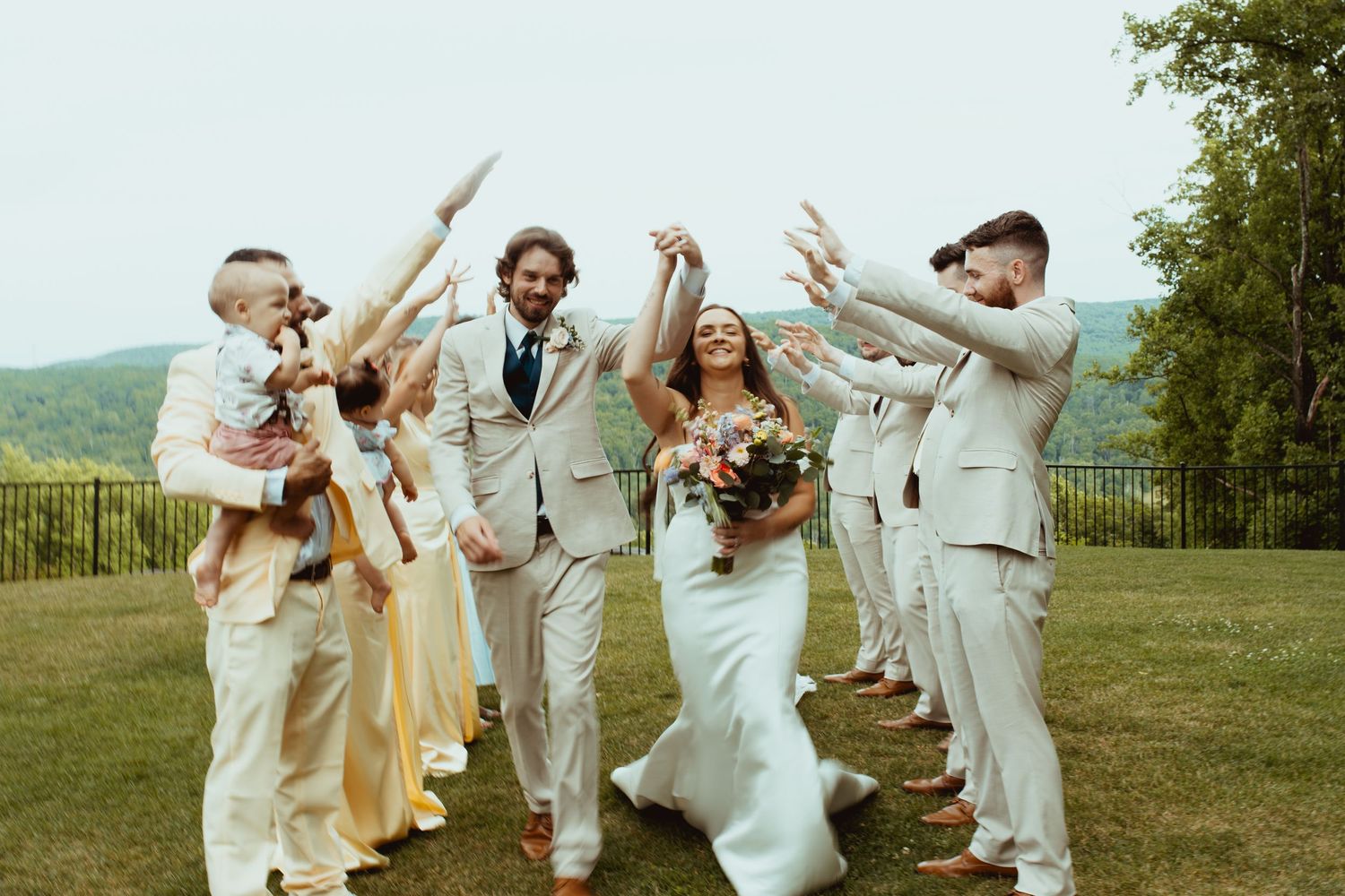 Joyful wedding party celebration outdoors, bride holding bouquet, groomsmen in tan suits, bridesmaids in yellow dresses, mountains behind.
