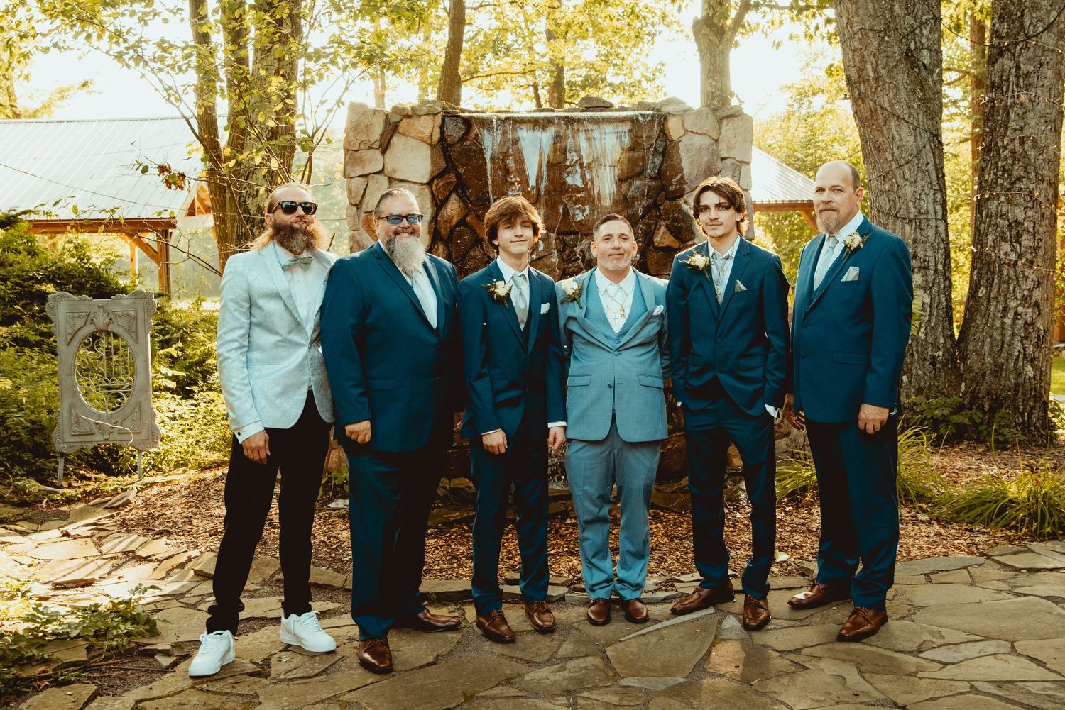 Wedding party in coordinated blue suits posing outdoors against stone wall.