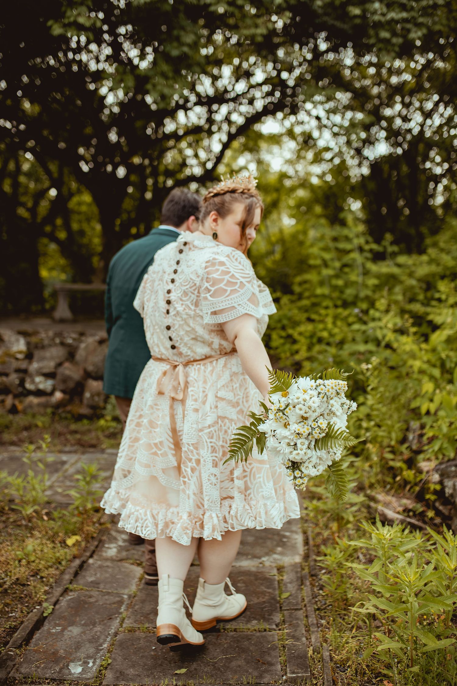 A small child in a white lace dress walks along a stone path surrounded by greenery.
