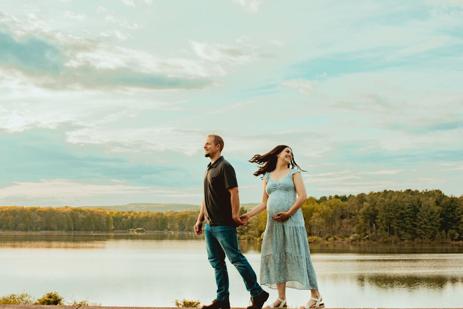 A couple walks along a serene lakeside path during sunset with a dog, creating a peaceful outdoor scene.