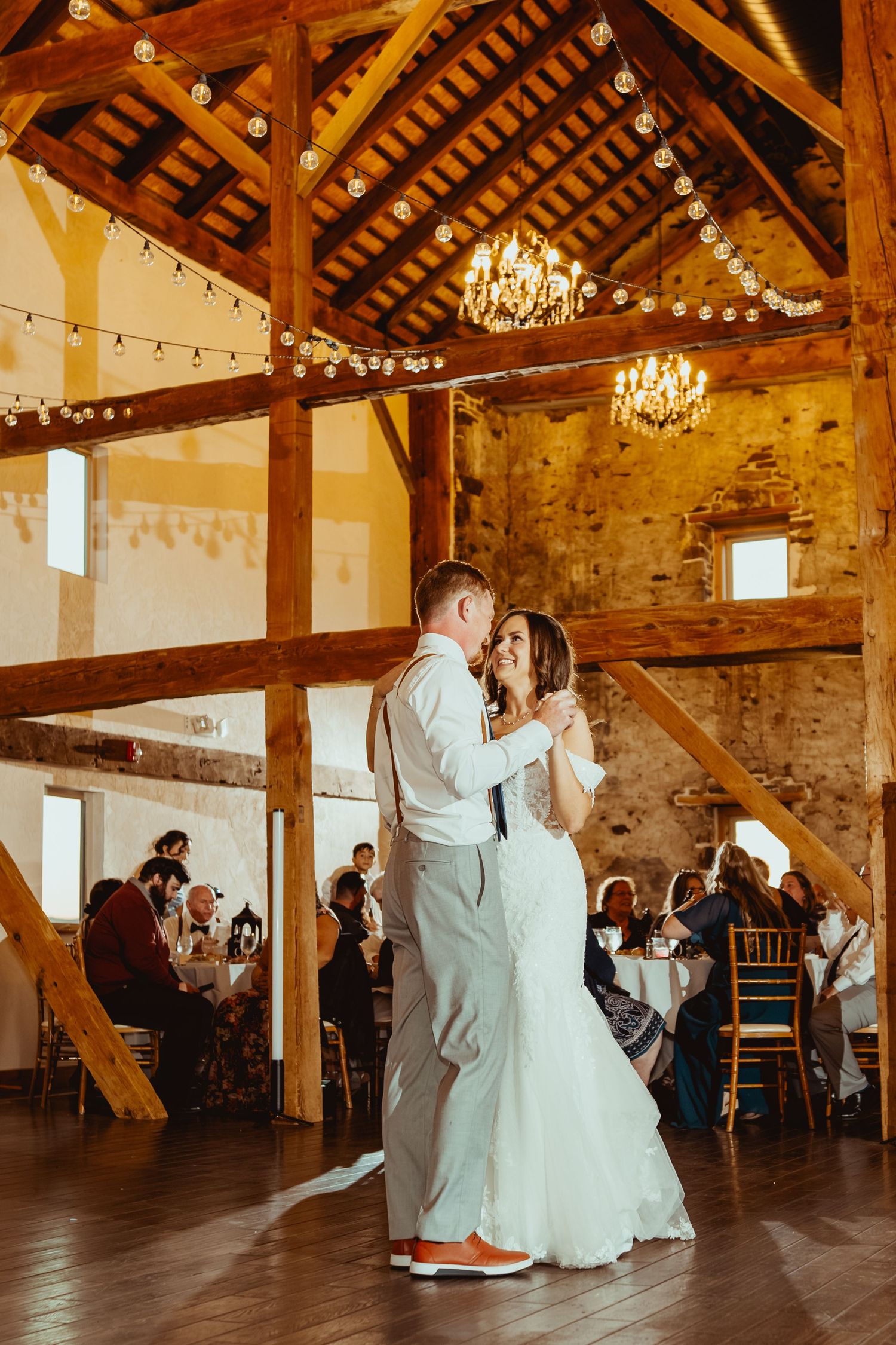 A couple dances together during their first dance in a rustic barn venue with exposed beams and chandeliers.
