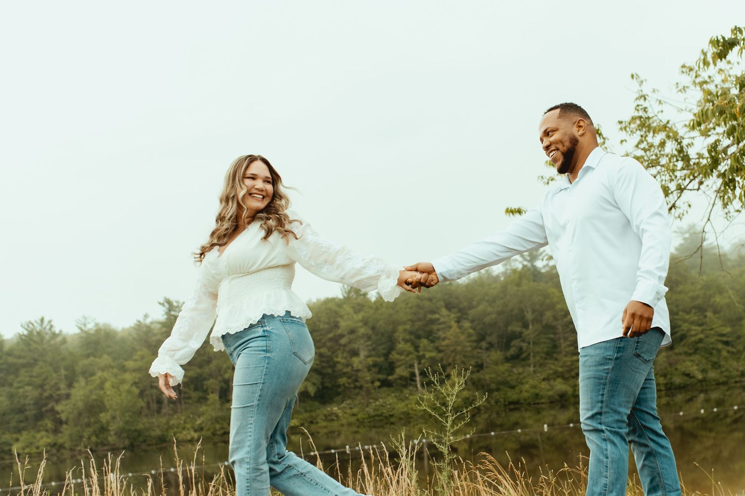 Happy couple holding hands and walking through a scenic field, wearing matching white tops and jeans on a cloudy day.
