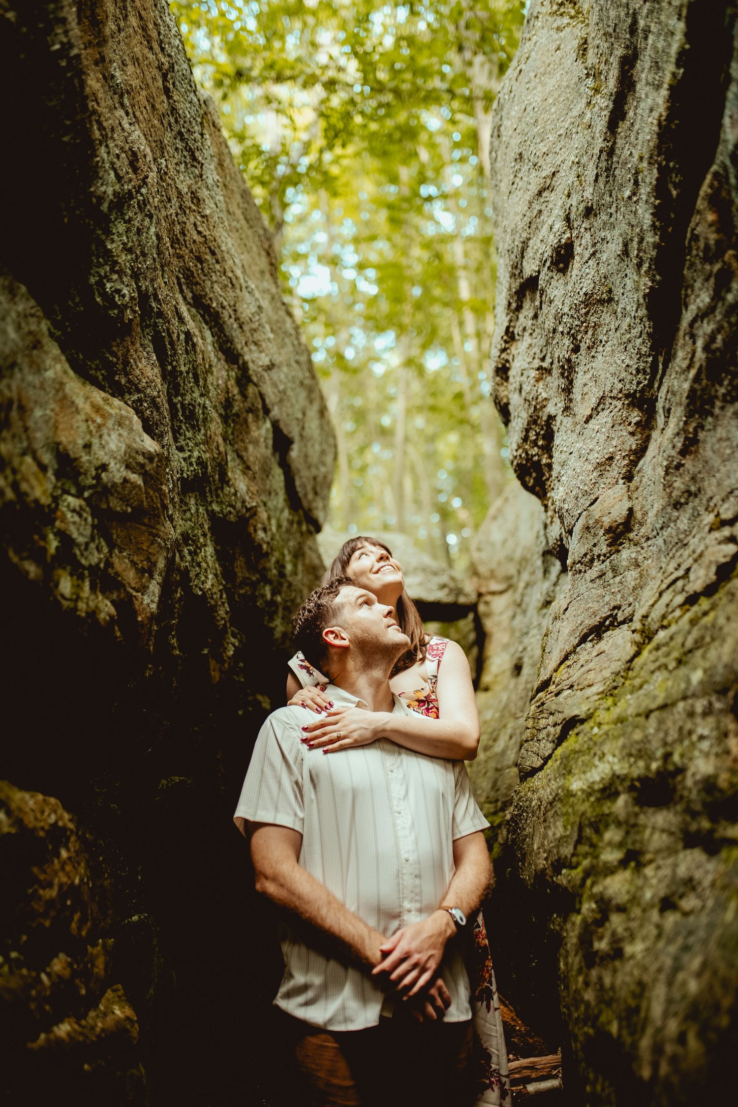 Couple embracing between tall rock formations in a lush forest, looking up with joy and wonder.