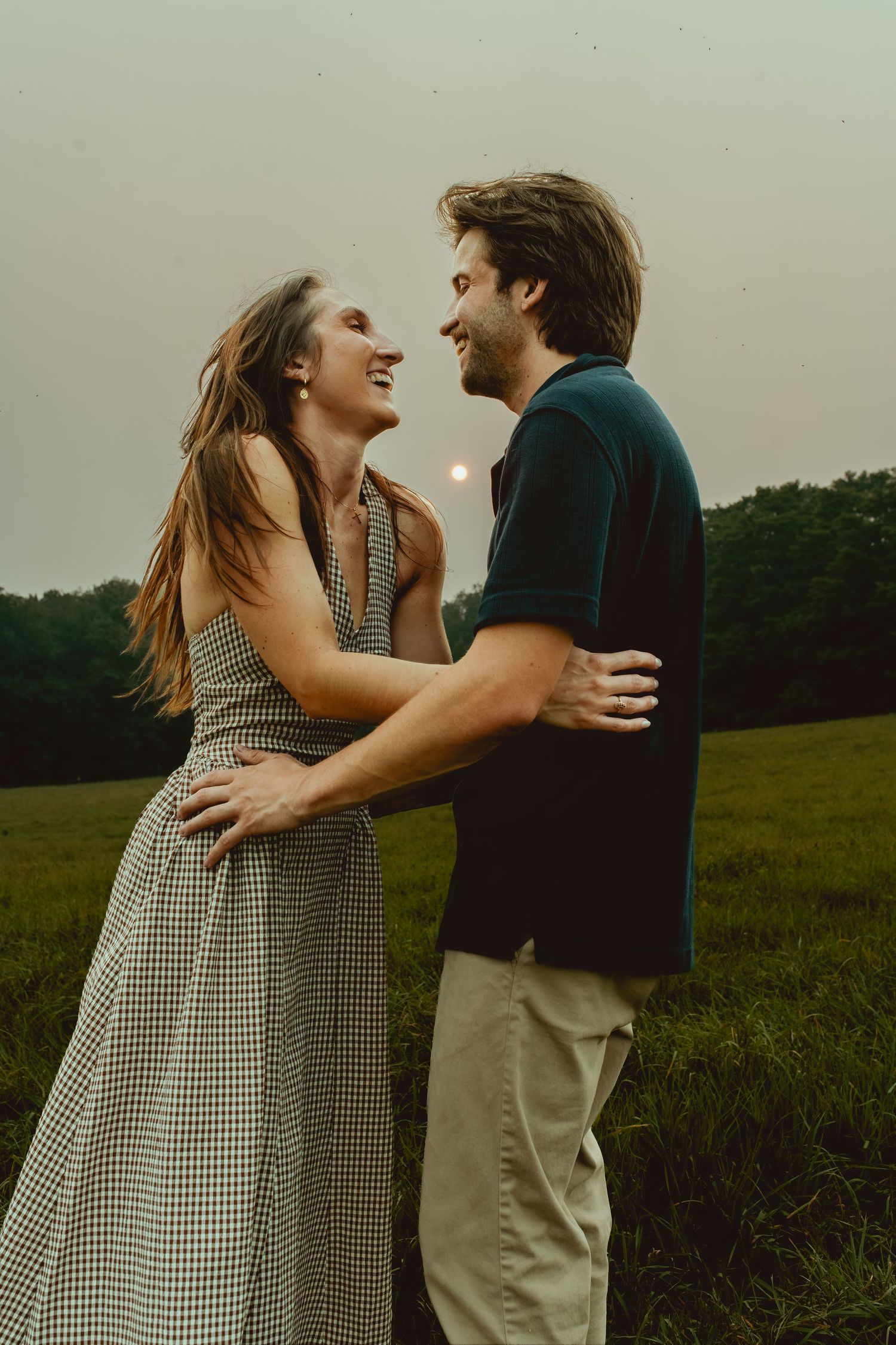 A joyful couple embraces and laughs together in a green field at dusk, woman in gingham dress, man in dark polo.