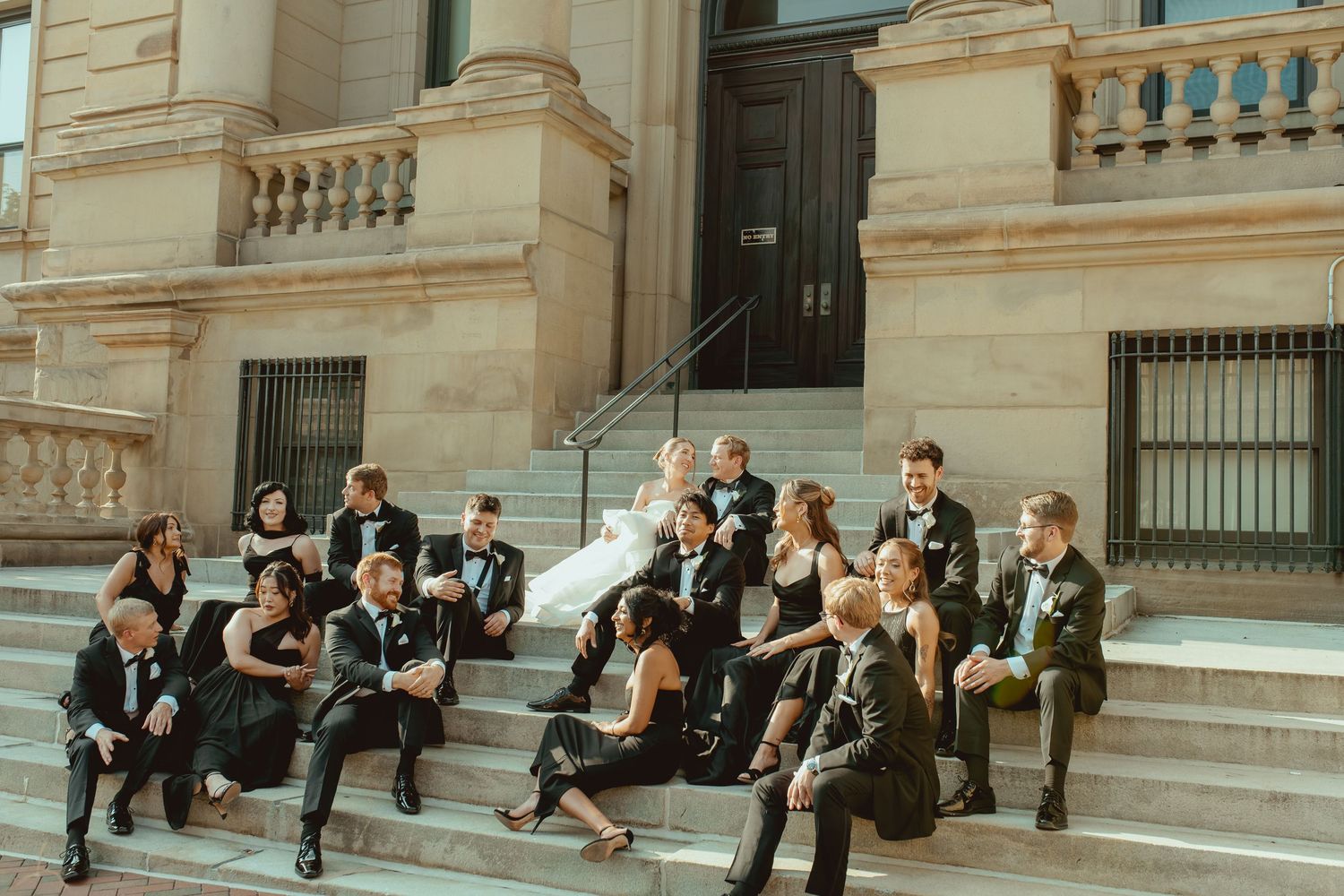 A group of formally dressed people sitting on stone steps of a grand building with classical architecture.