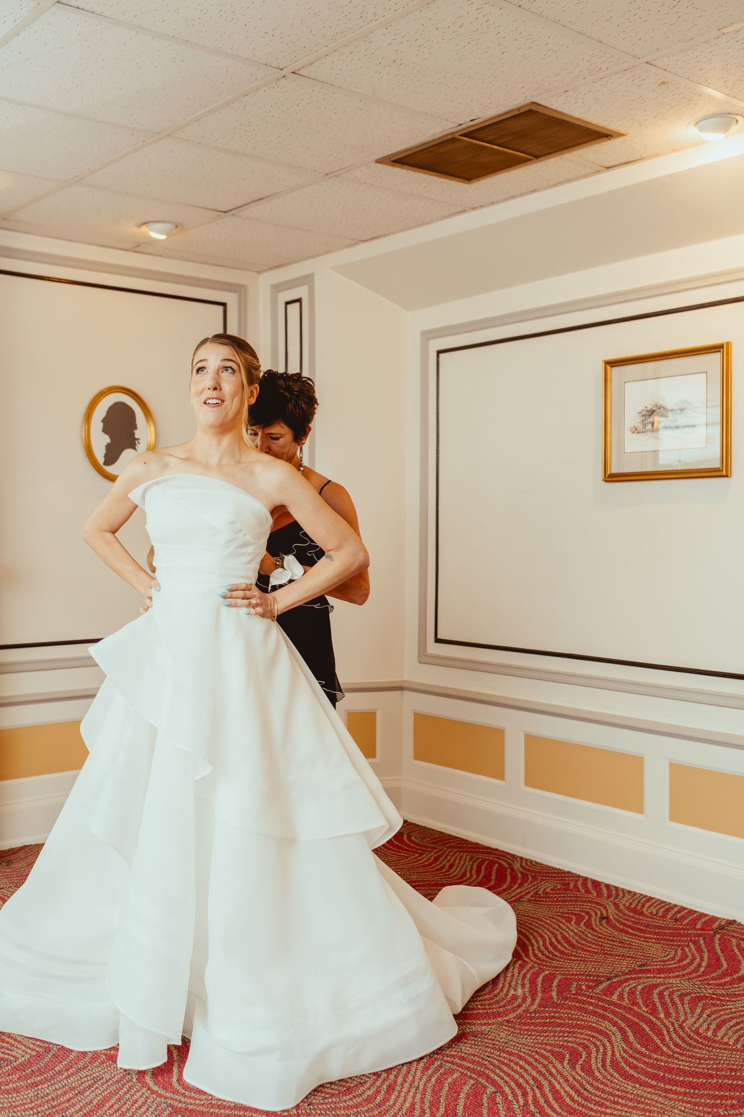 A stunning white wedding gown being fitted in an elegant room with yellow accents and a patterned red carpet.