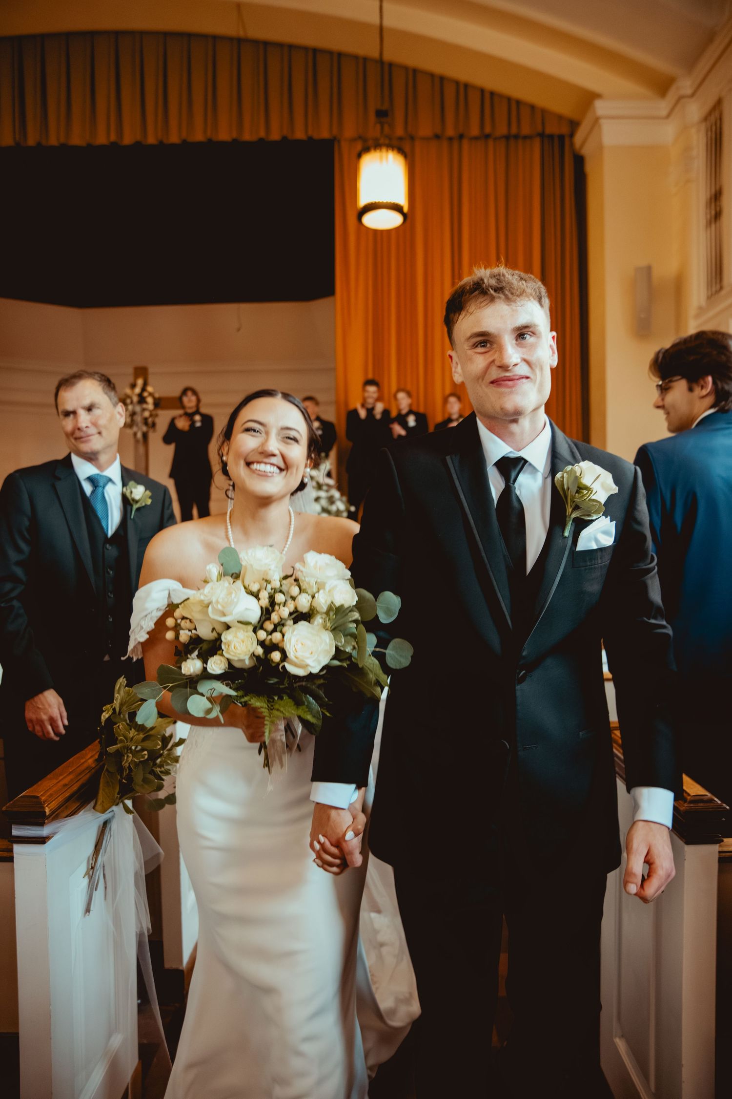 Wedding party poses in formal attire with white bouquets against warm interior lighting.