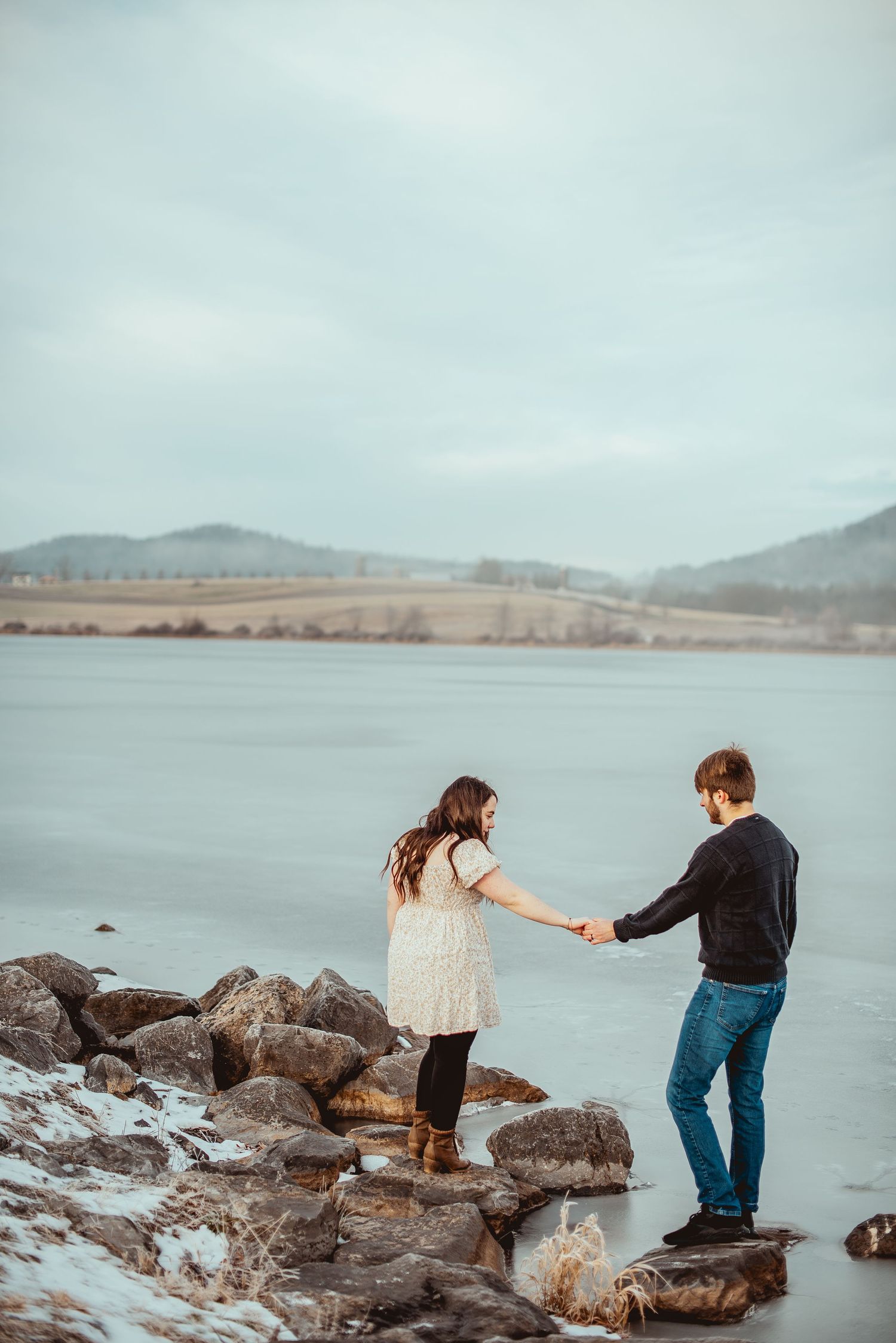 Two people explore a rocky shoreline along a misty lake with mountains in the background.