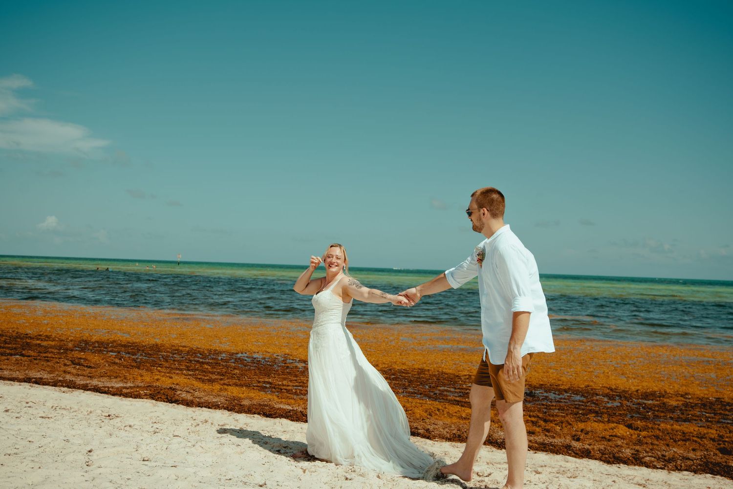 Bride and groom hold hands walking on a tropical beach, with turquoise water and seaweed shoreline in the background.
