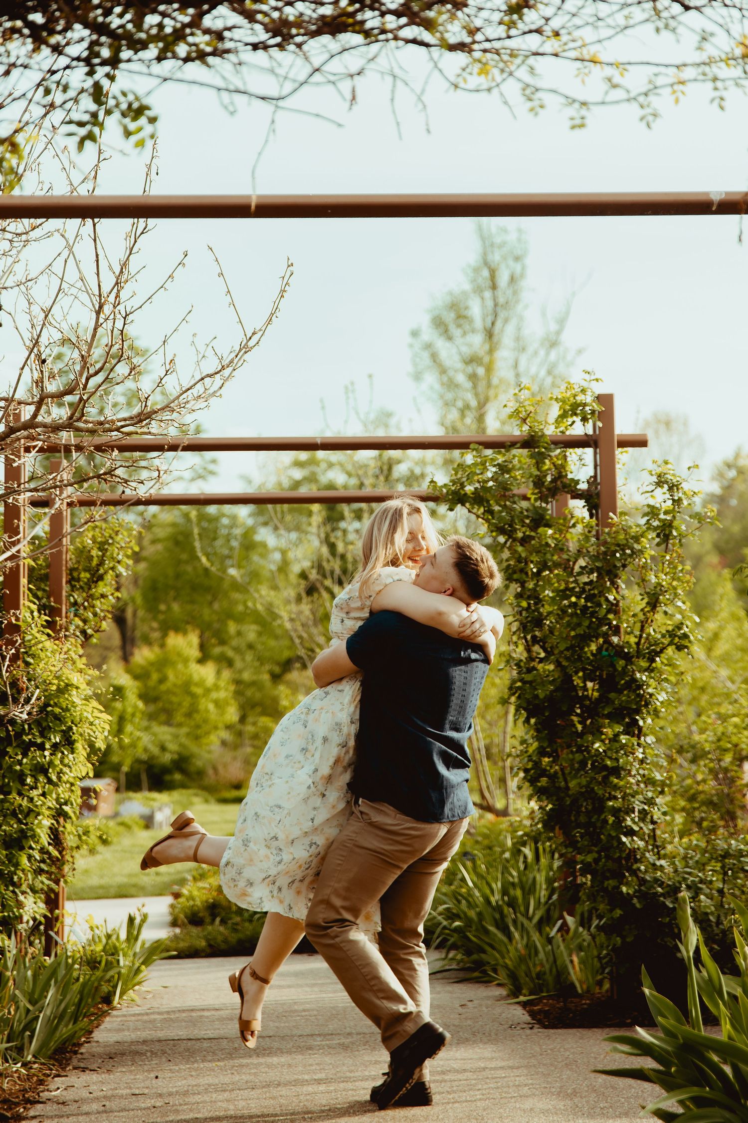 Couple embracing and spinning joyfully on a garden path surrounded by lush greenery and a wooden pergola.