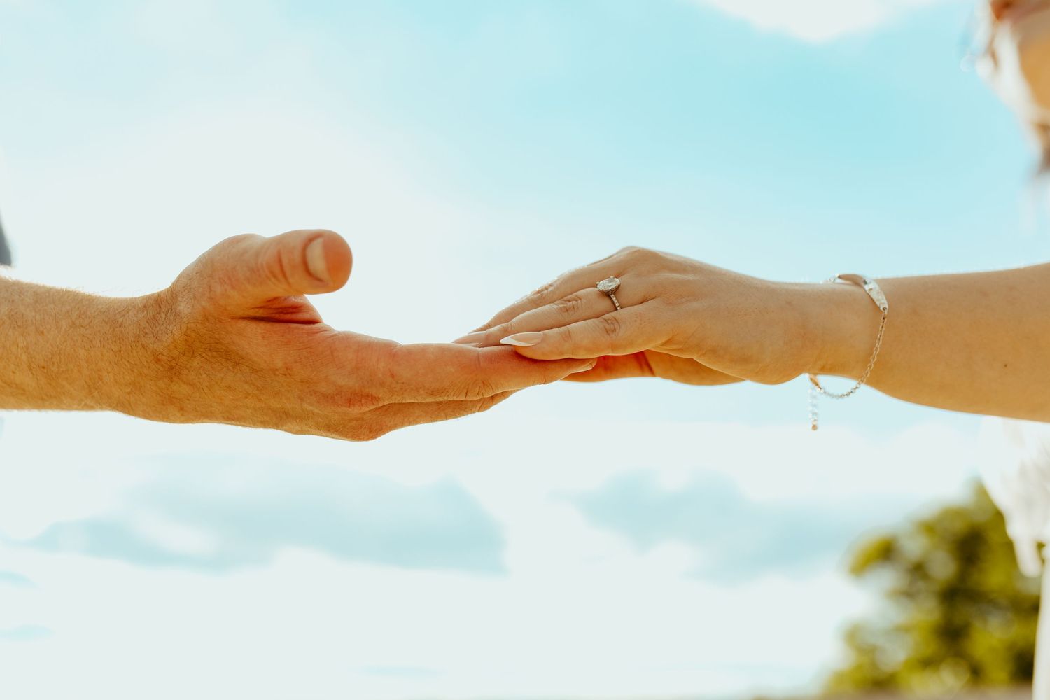 Two hands gently touching against a blue sky, woman wearing a diamond ring and delicate bracelet.