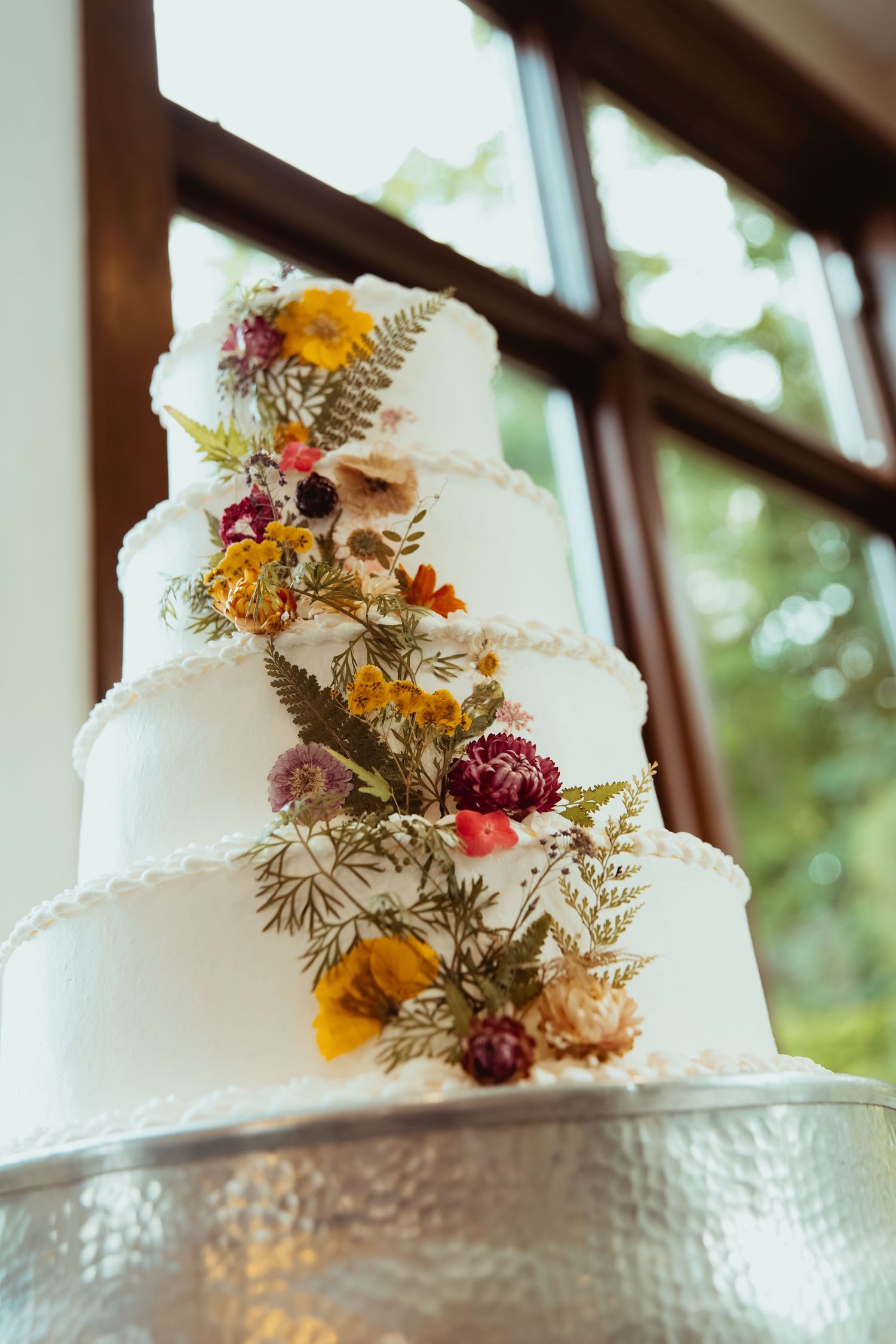 Elegant four-tier white wedding cake decorated with colorful dried flowers and ferns near a sunlit window.