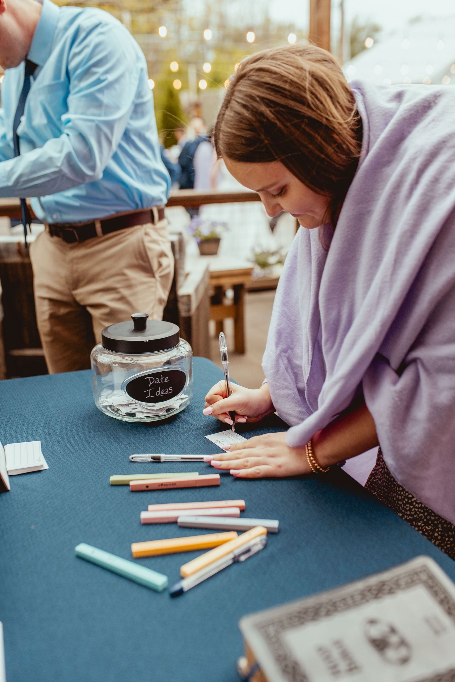 Wedding guest writing date night ideas on card at reception table with colorful markers and glass jar.