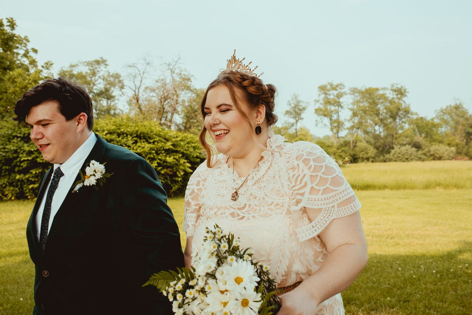 A joyful wedding couple walks together outdoors in a sunny field holding a white floral bouquet.