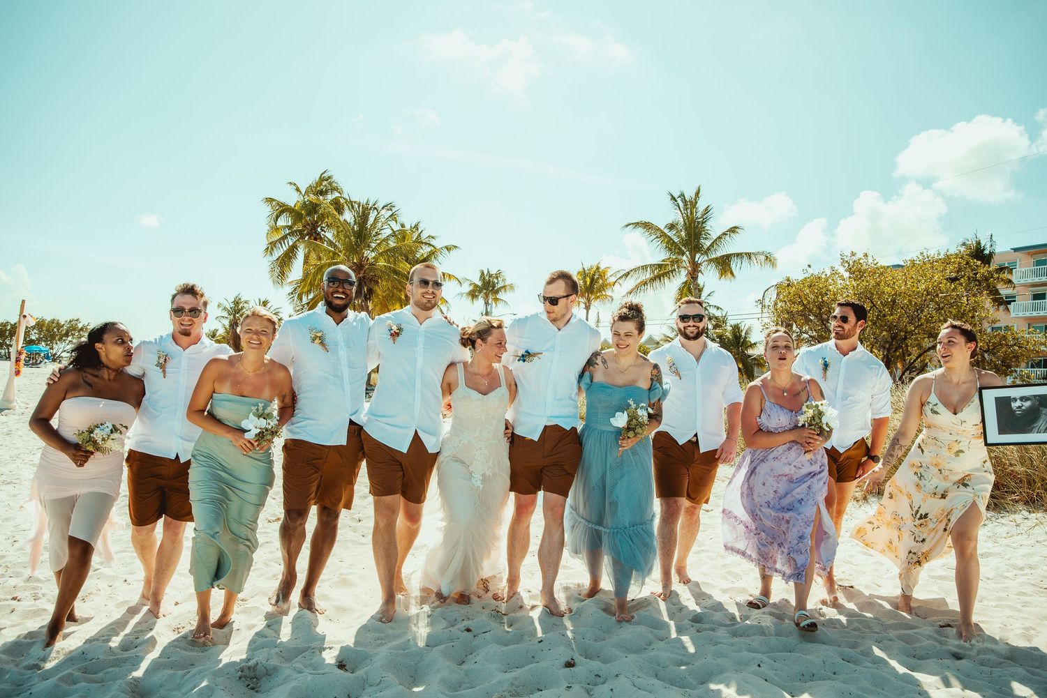 Wedding party celebrating on a tropical beach with palm trees, wearing casual attire in blue and white tones.