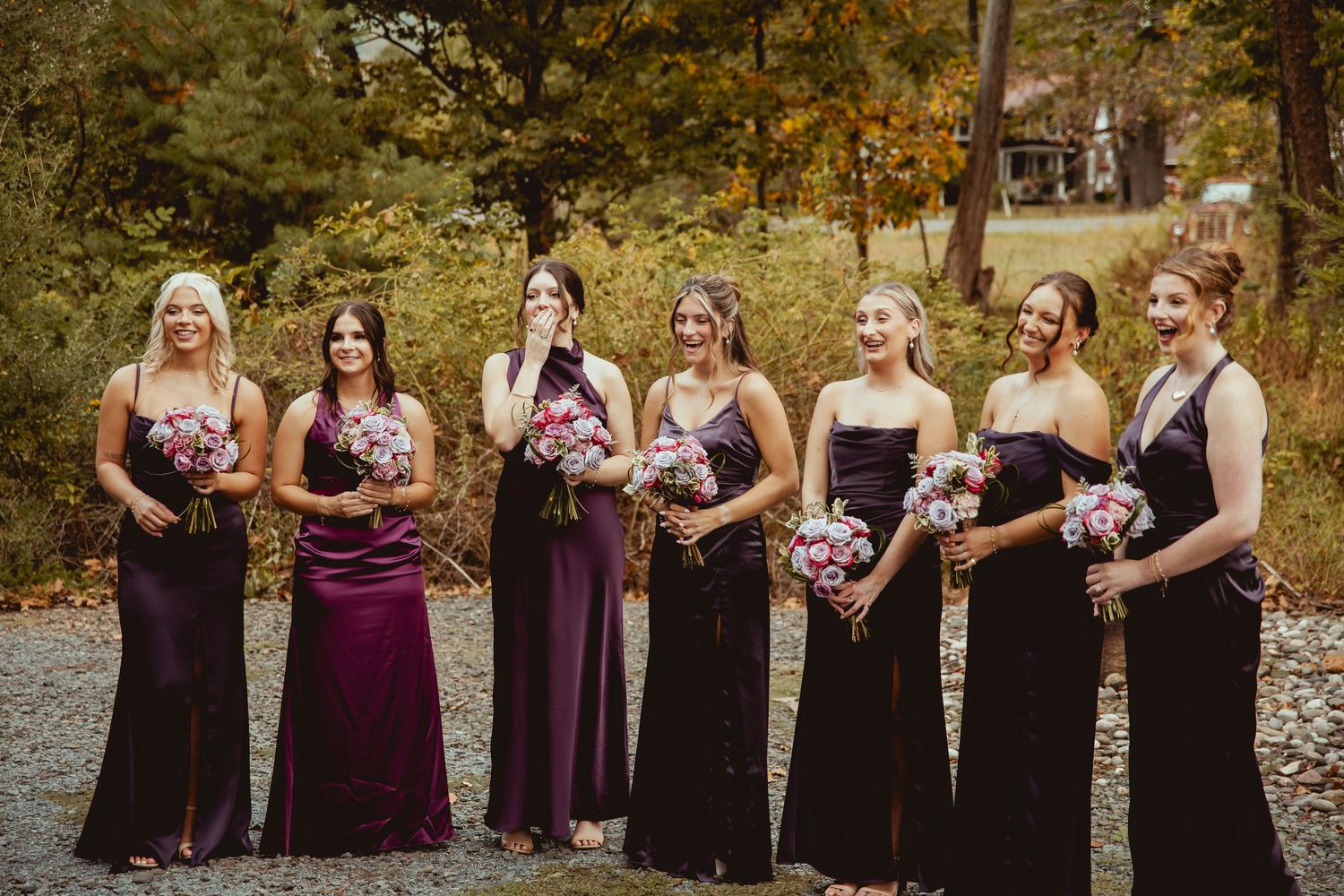 Bridesmaids in burgundy and black dresses stand together in a row during an autumn outdoor wedding photoshoot.