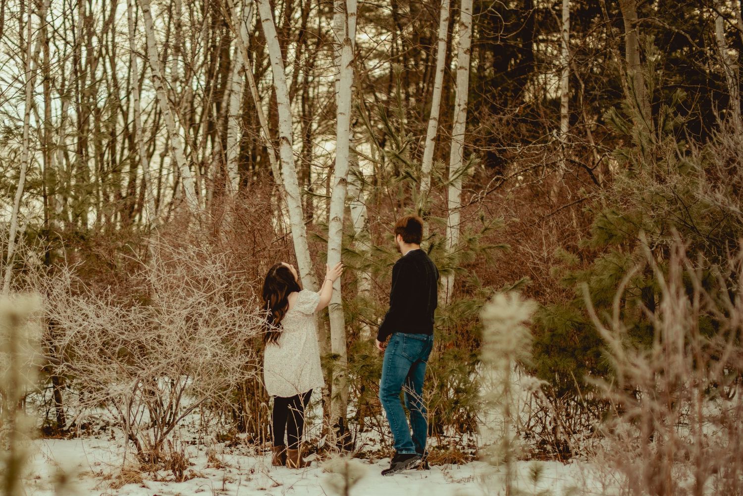 A person stands in a snowy winter forest setting with bare trees and white ground cover.