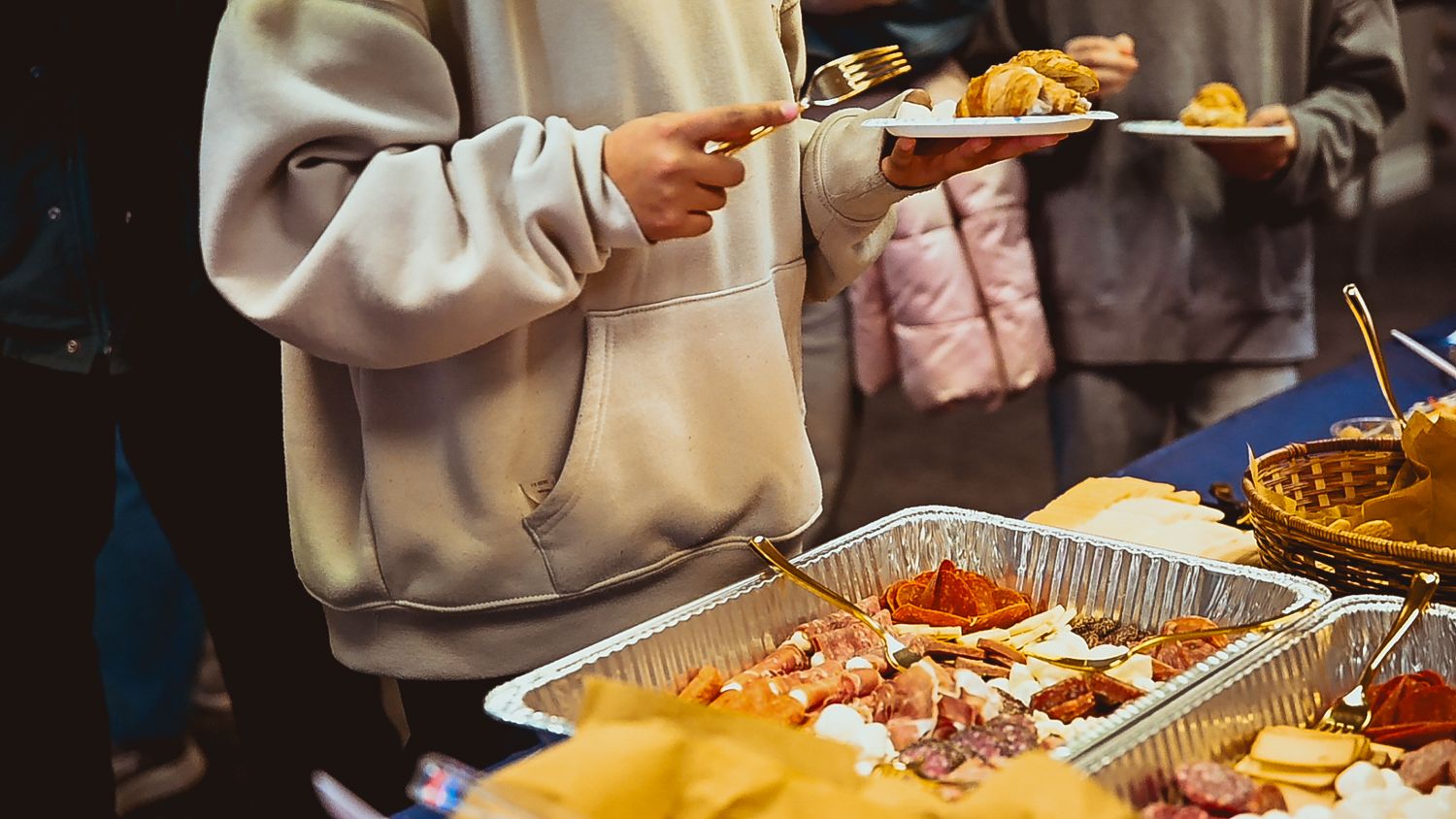 Someone in a beige sweatshirt serving food from aluminum catering trays at a buffet-style setup.