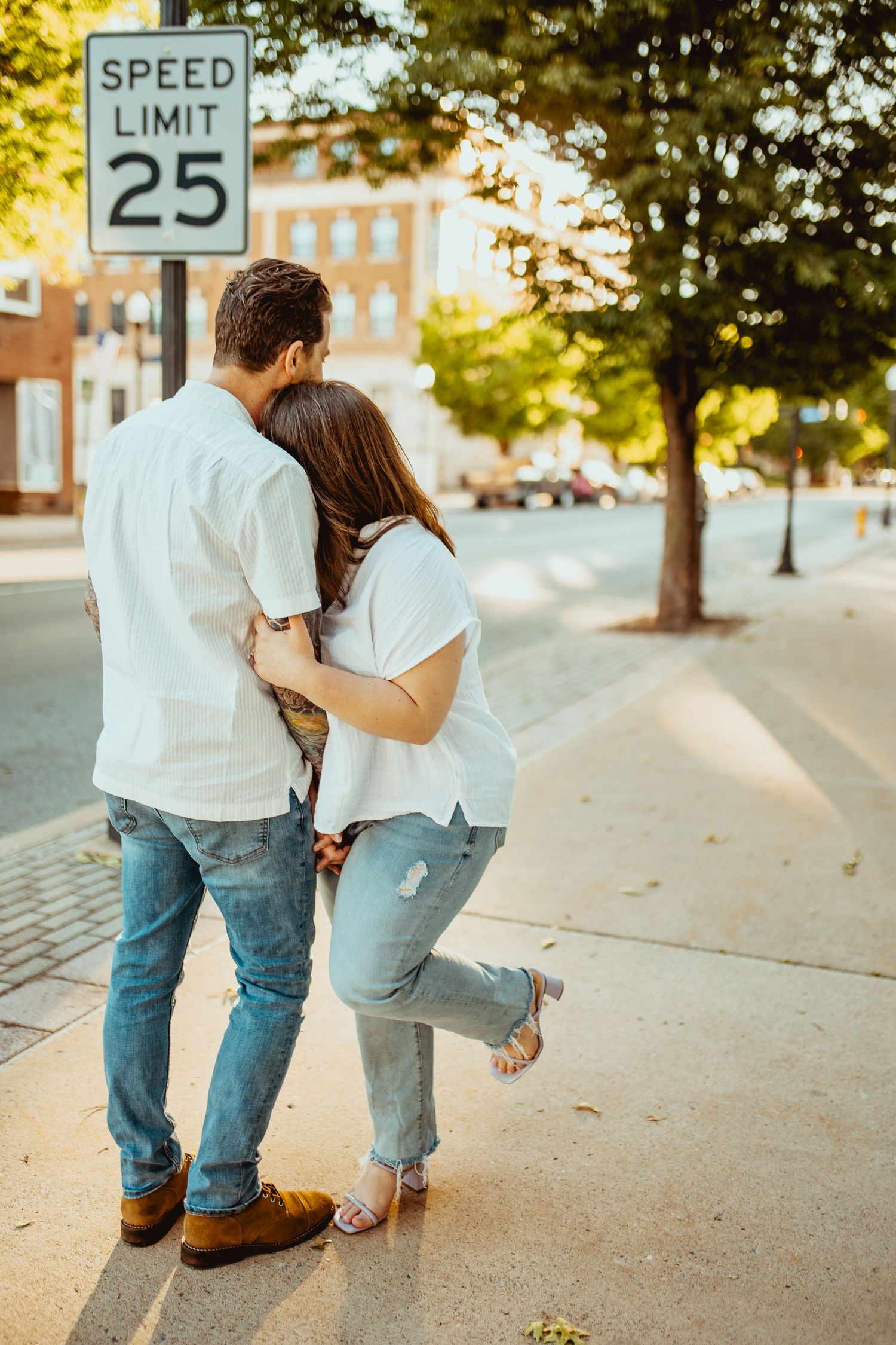 A couple hugging near a speed limit 25 sign on a sunny urban sidewalk surrounded by trees.