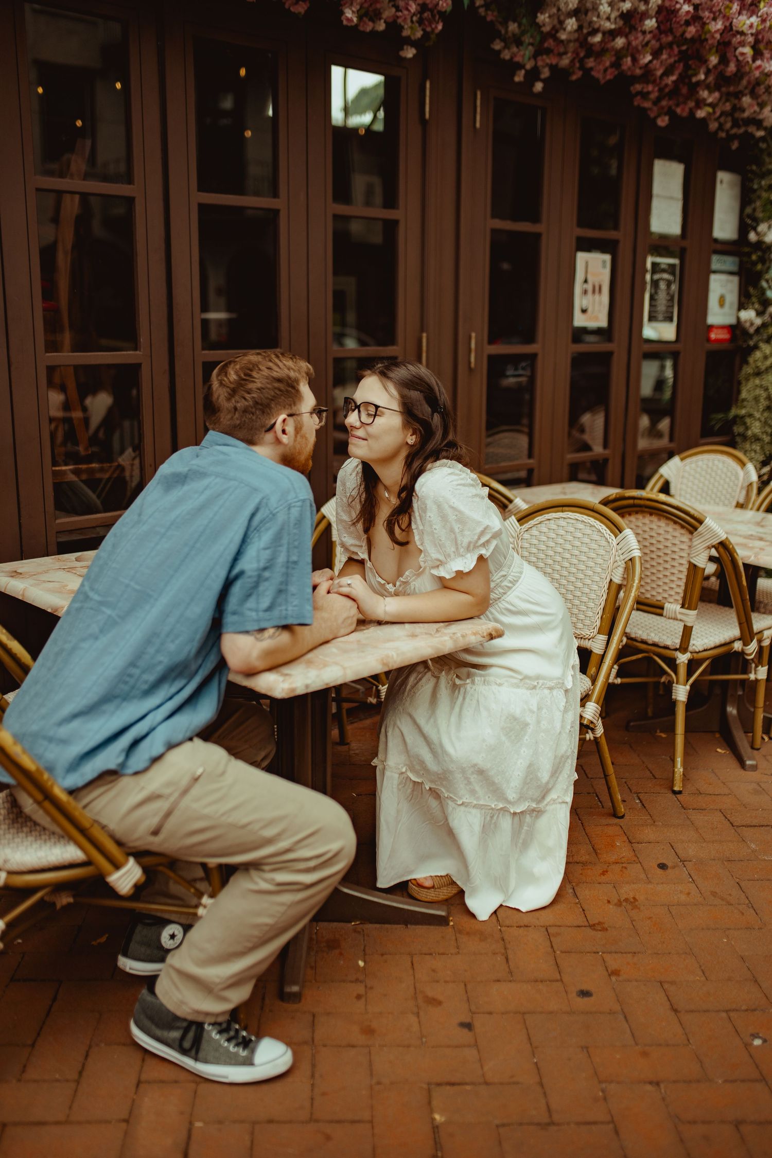 Couple holding hands at outdoor cafe table, woman in white dress and man in blue shirt sharing an intimate moment.