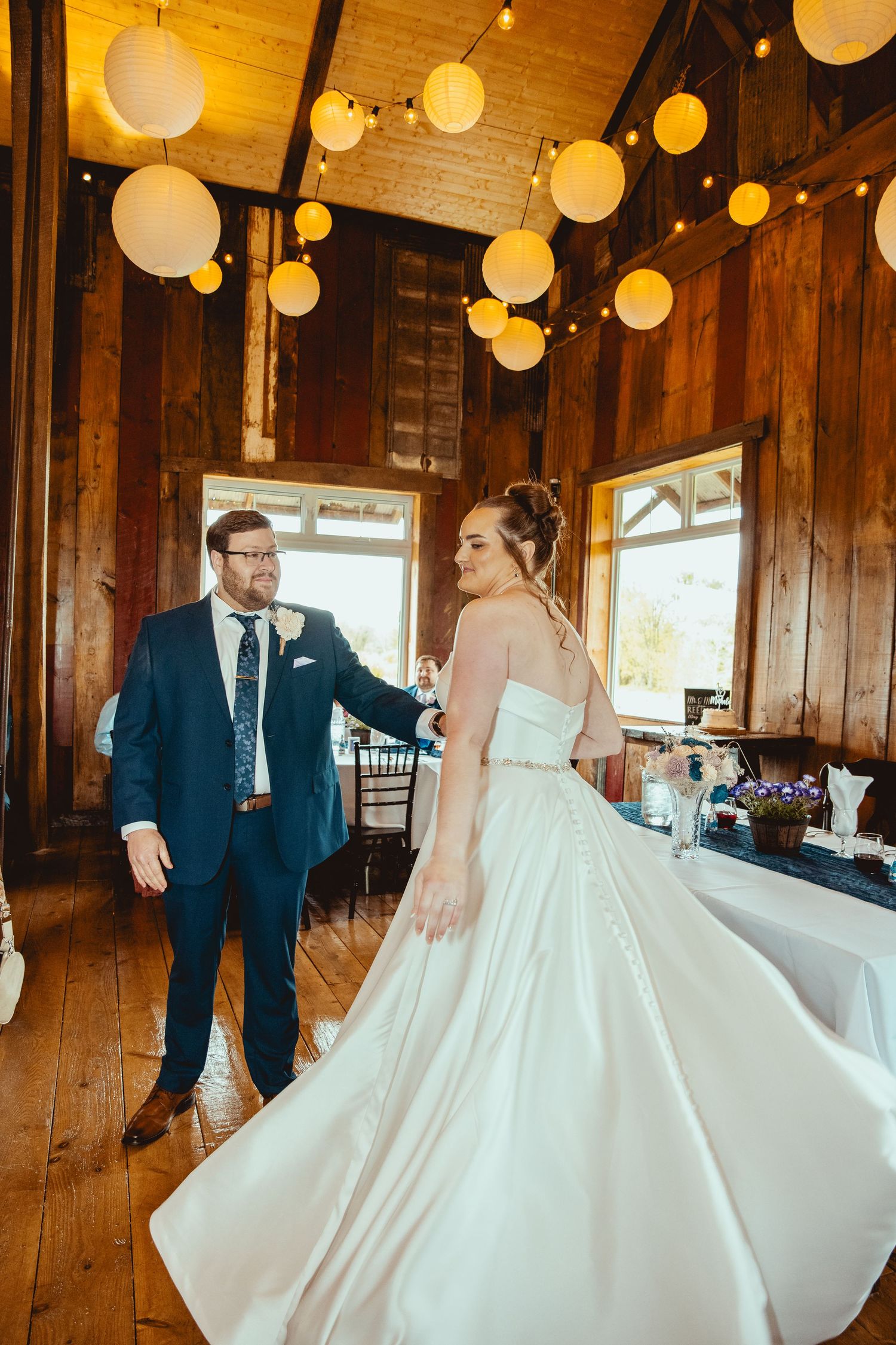 Bride in strapless white ball gown dancing with groom in navy suit inside rustic barn with string lights.