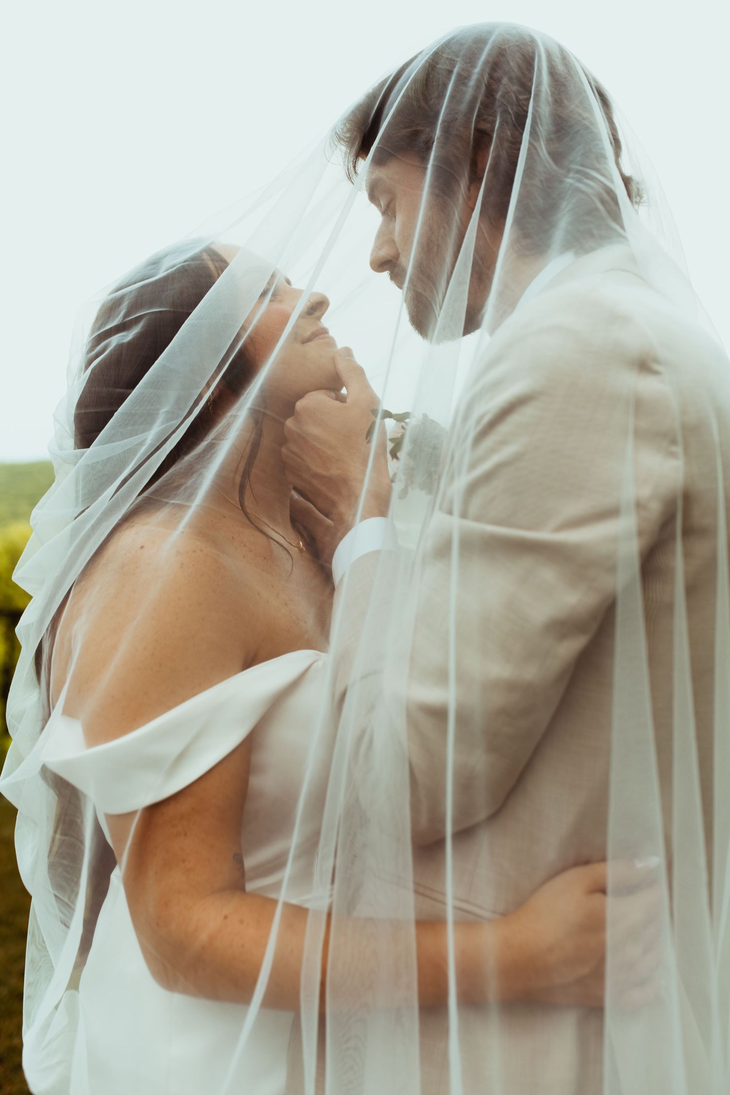Bride and groom share intimate moment together under flowing white veil at outdoor wedding.