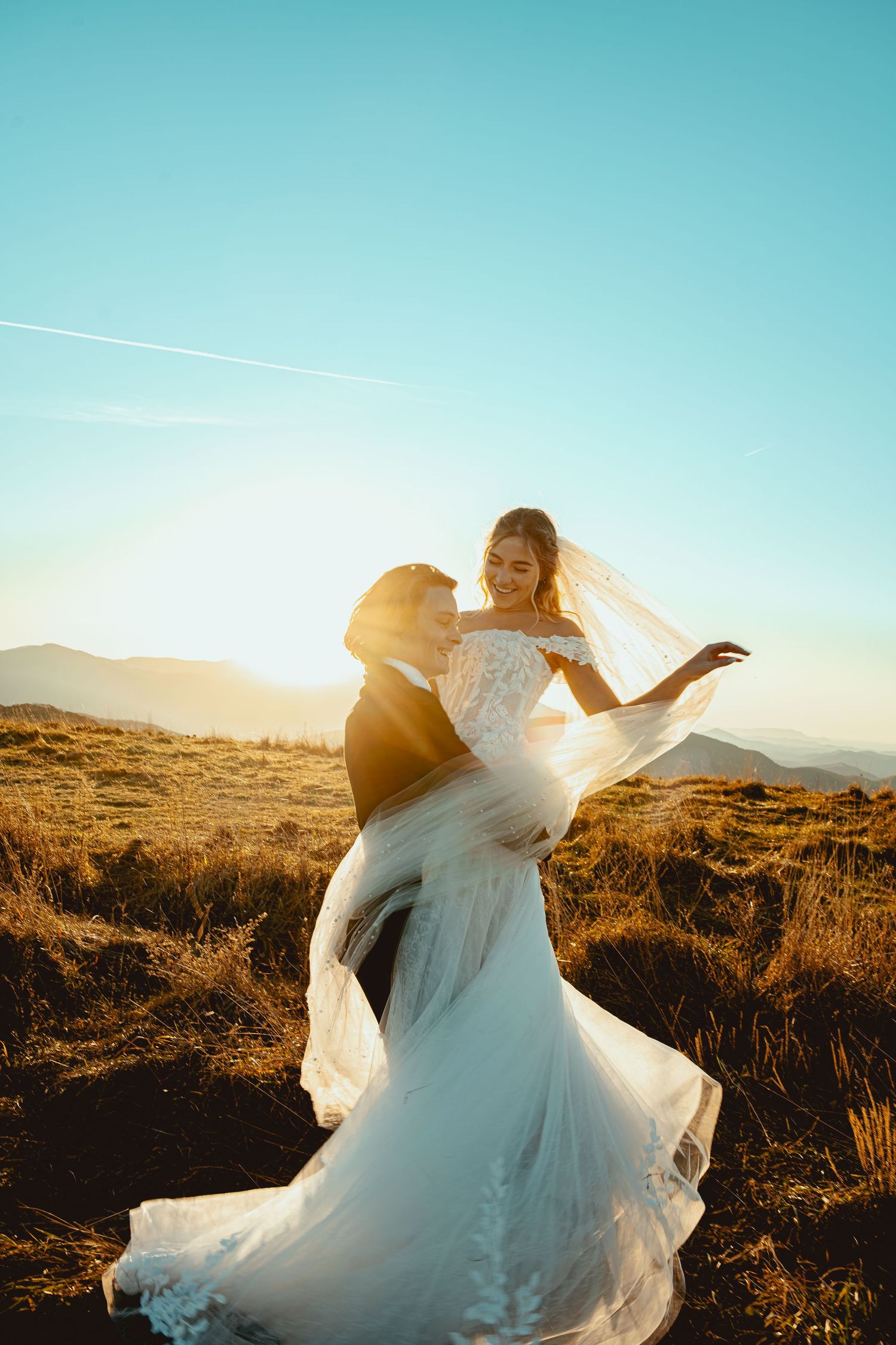 Bride in white gown held by groom at golden sunset on mountain meadow, veil flowing in wind.
