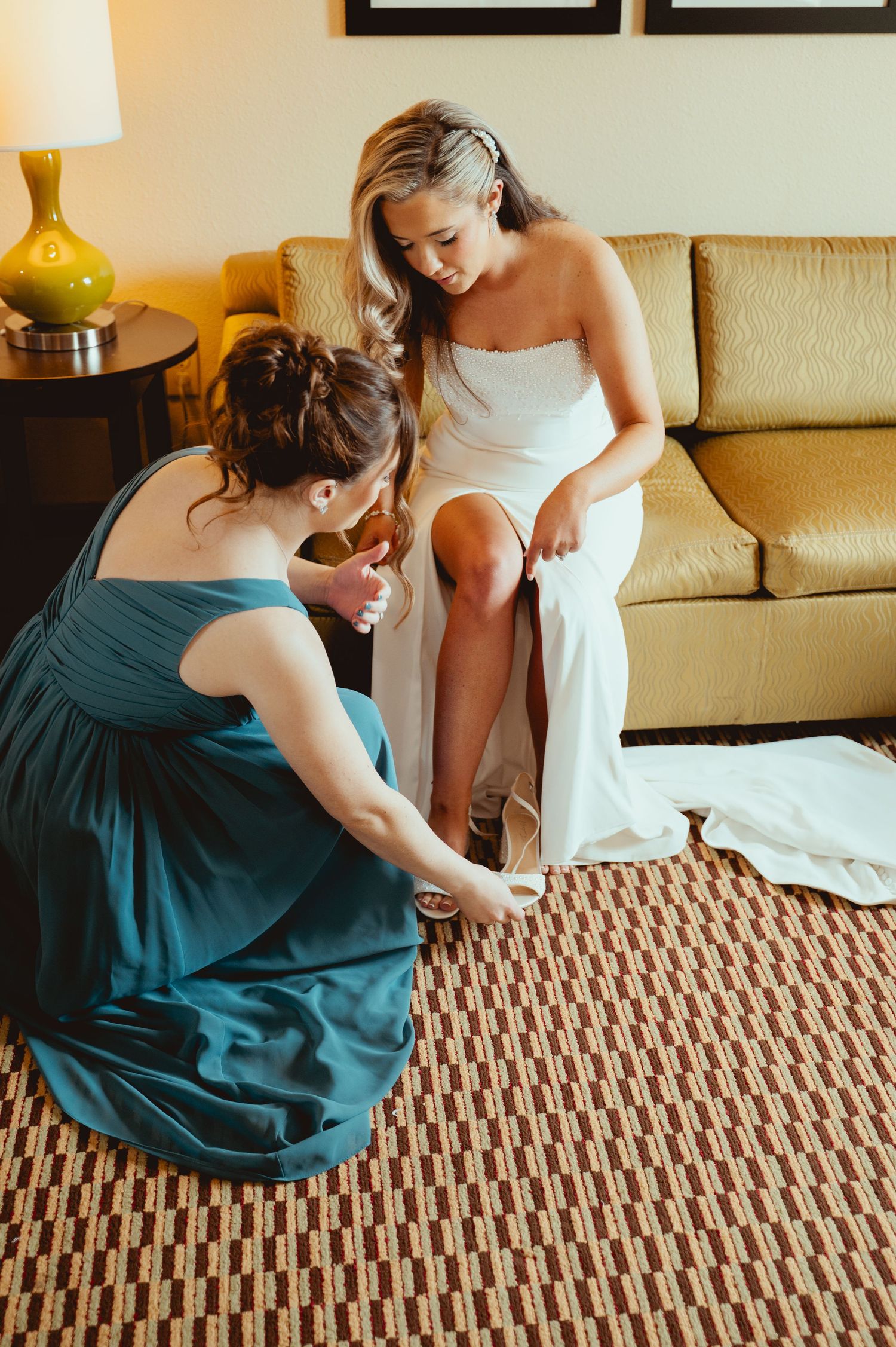 Bridesmaid in teal dress helps bride in white gown put on heels while sitting on hotel room sofa.