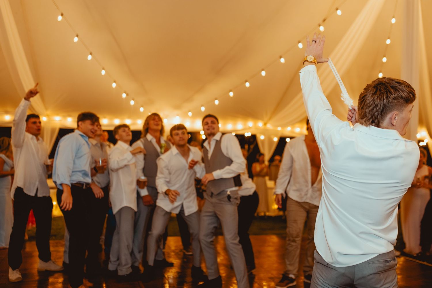 Groomsmen and guests celebrate energetically on dance floor under warm string lights at wedding reception.