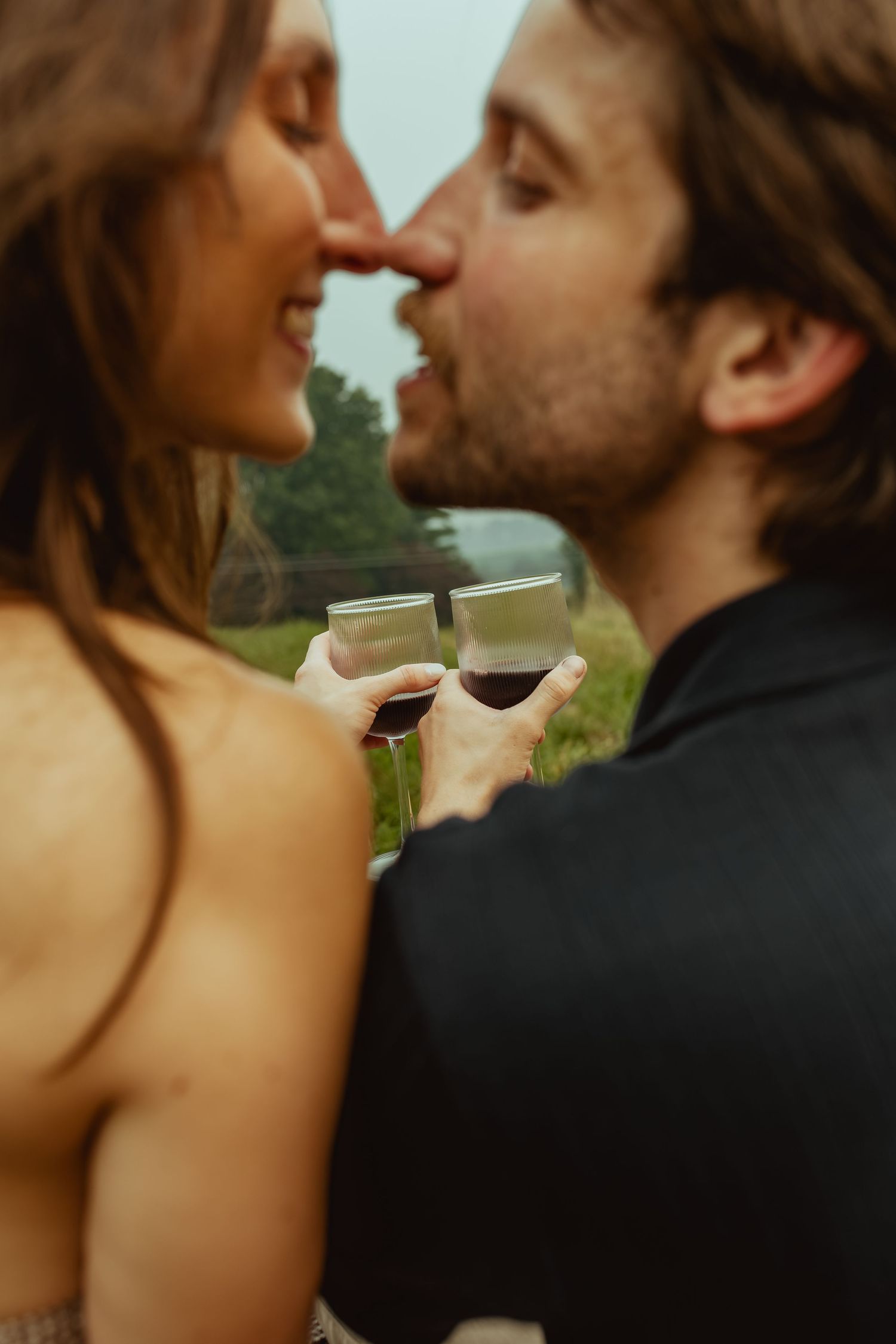 Romantic couple sharing a kiss while clinking glasses of red wine outdoors in a lush green setting.