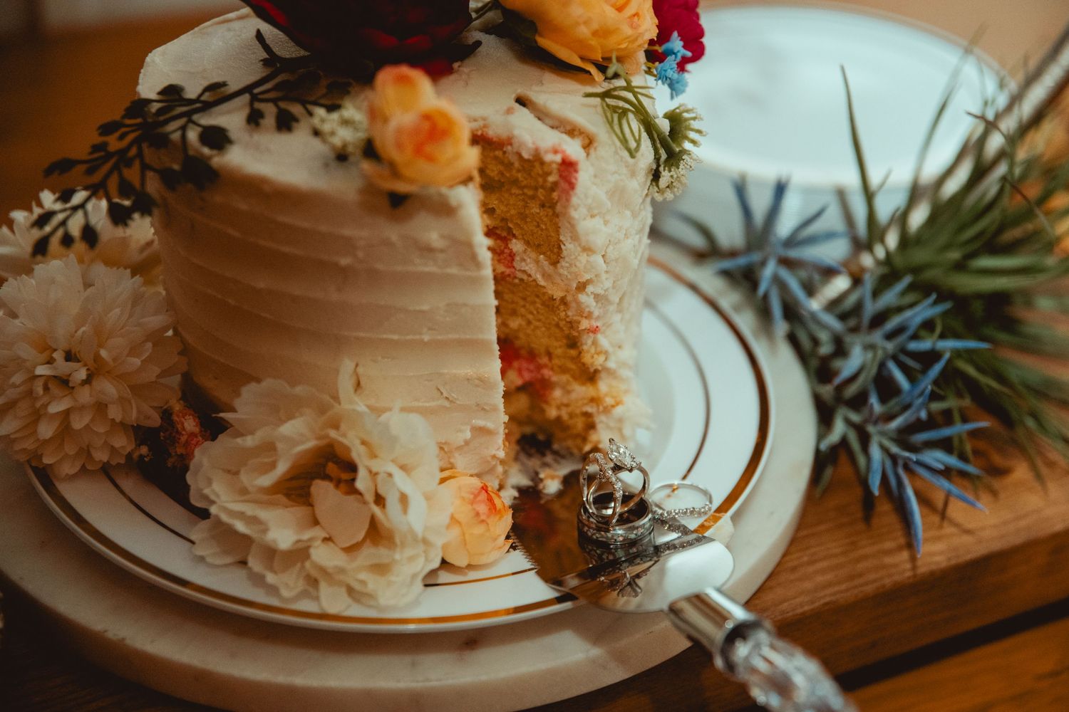 Rustic wedding cake with white frosting and colorful floral decorations, partially sliced on a decorative plate.