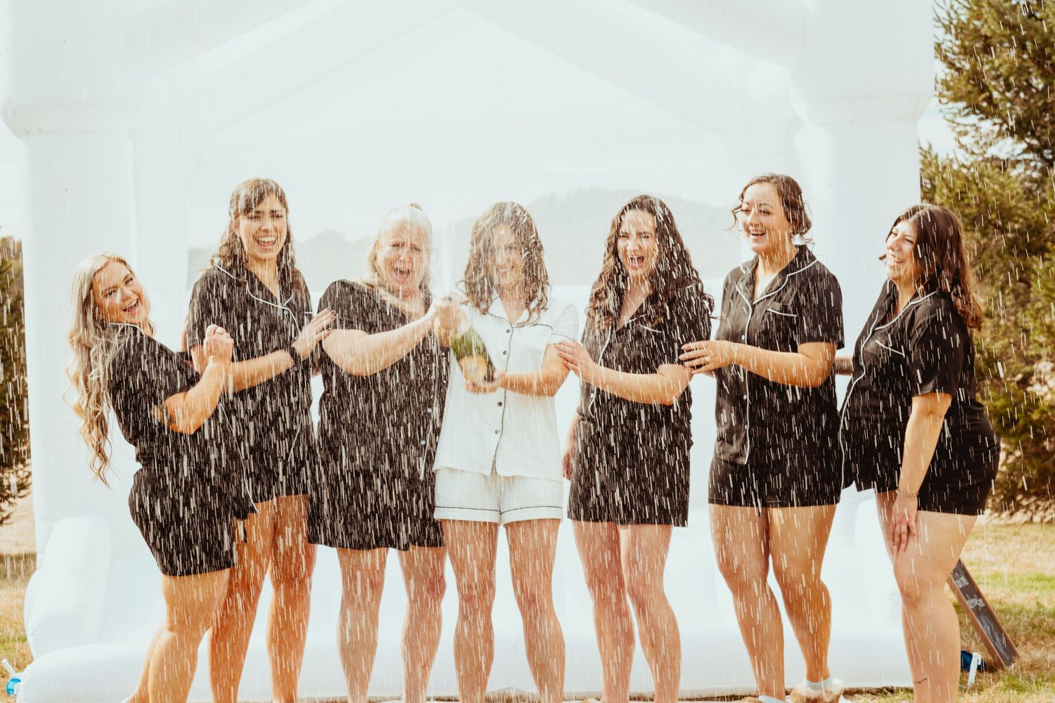 Bridal party celebrating with champagne sprays outdoors on a sunny day.