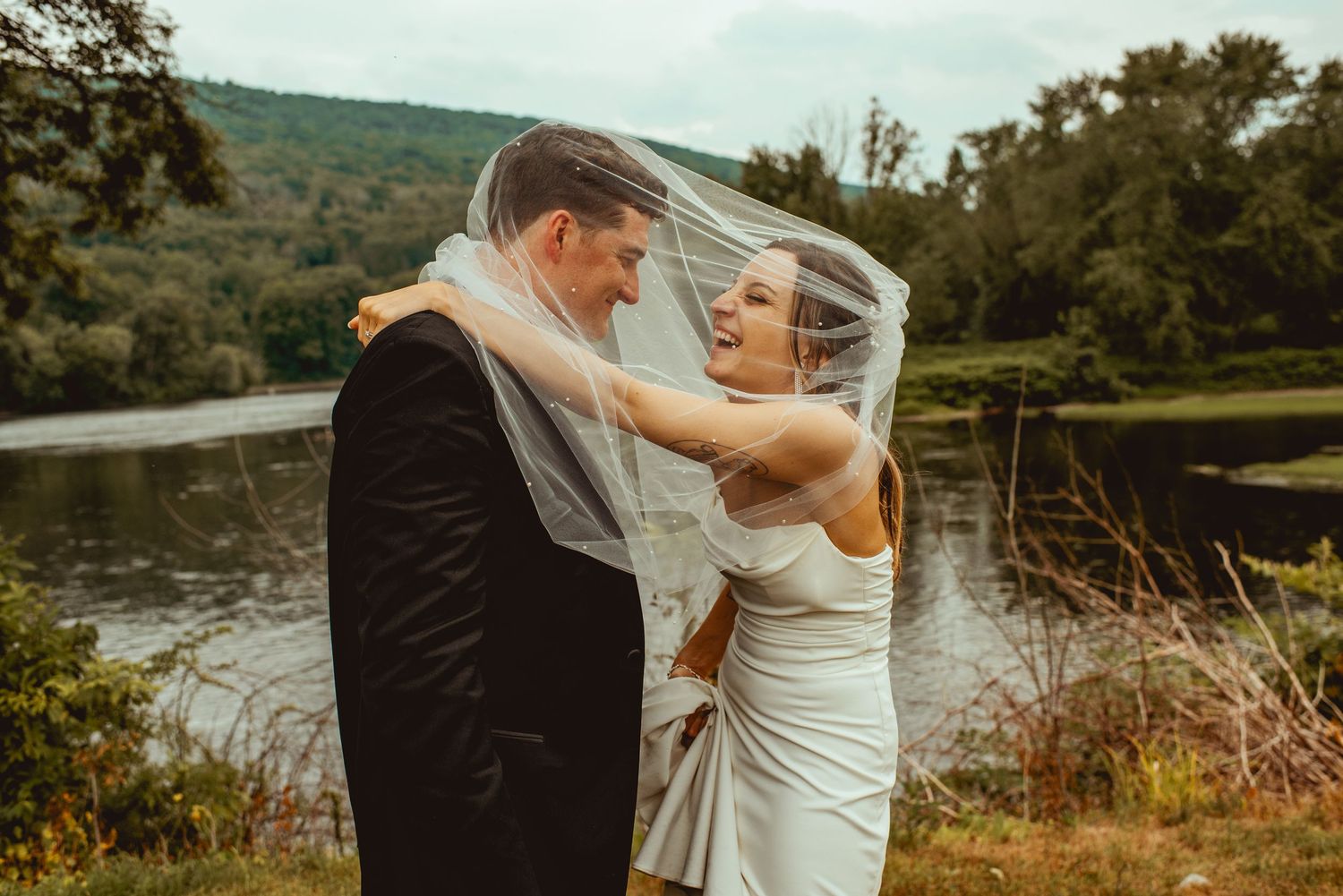 A romantic couple embraces near a scenic lake during their outdoor wedding celebration at sunset.