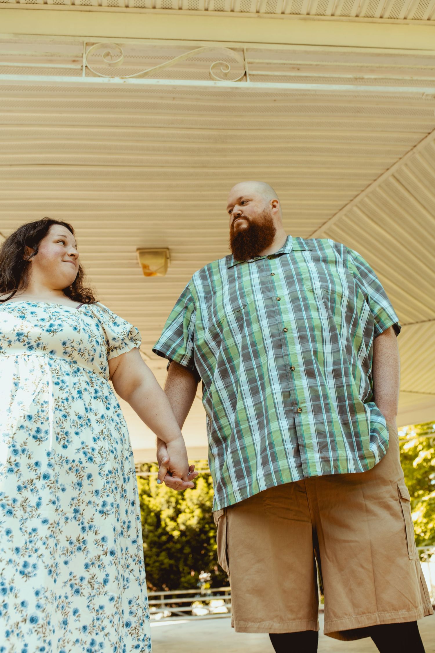 A plus-size couple holds hands and smiles at each other under a porch, wearing casual summer outfits.