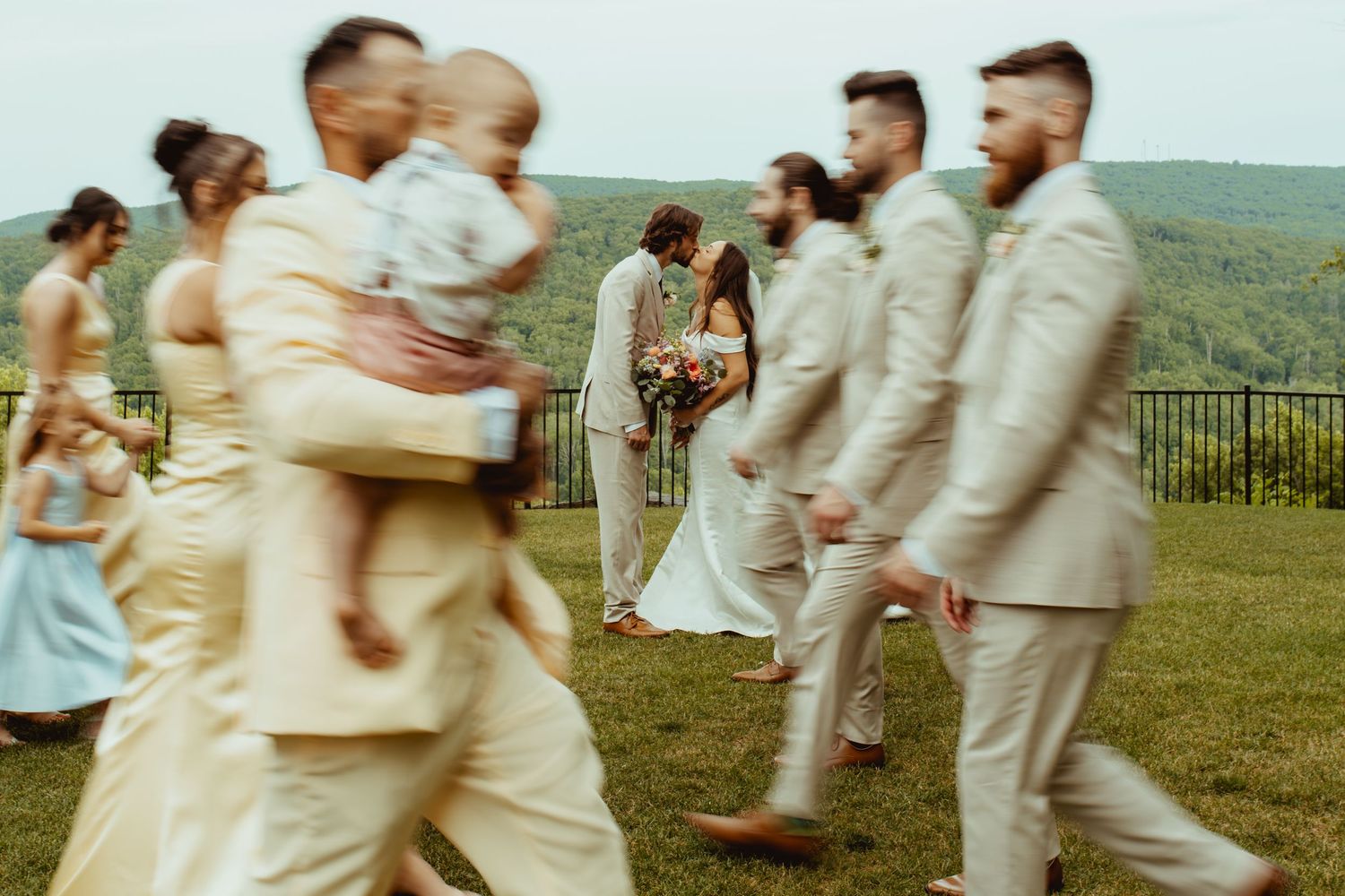 Wedding party walks outdoors on green hillside, groomsmen in tan suits and bridesmaids in yellow dresses, bride and groom in background.