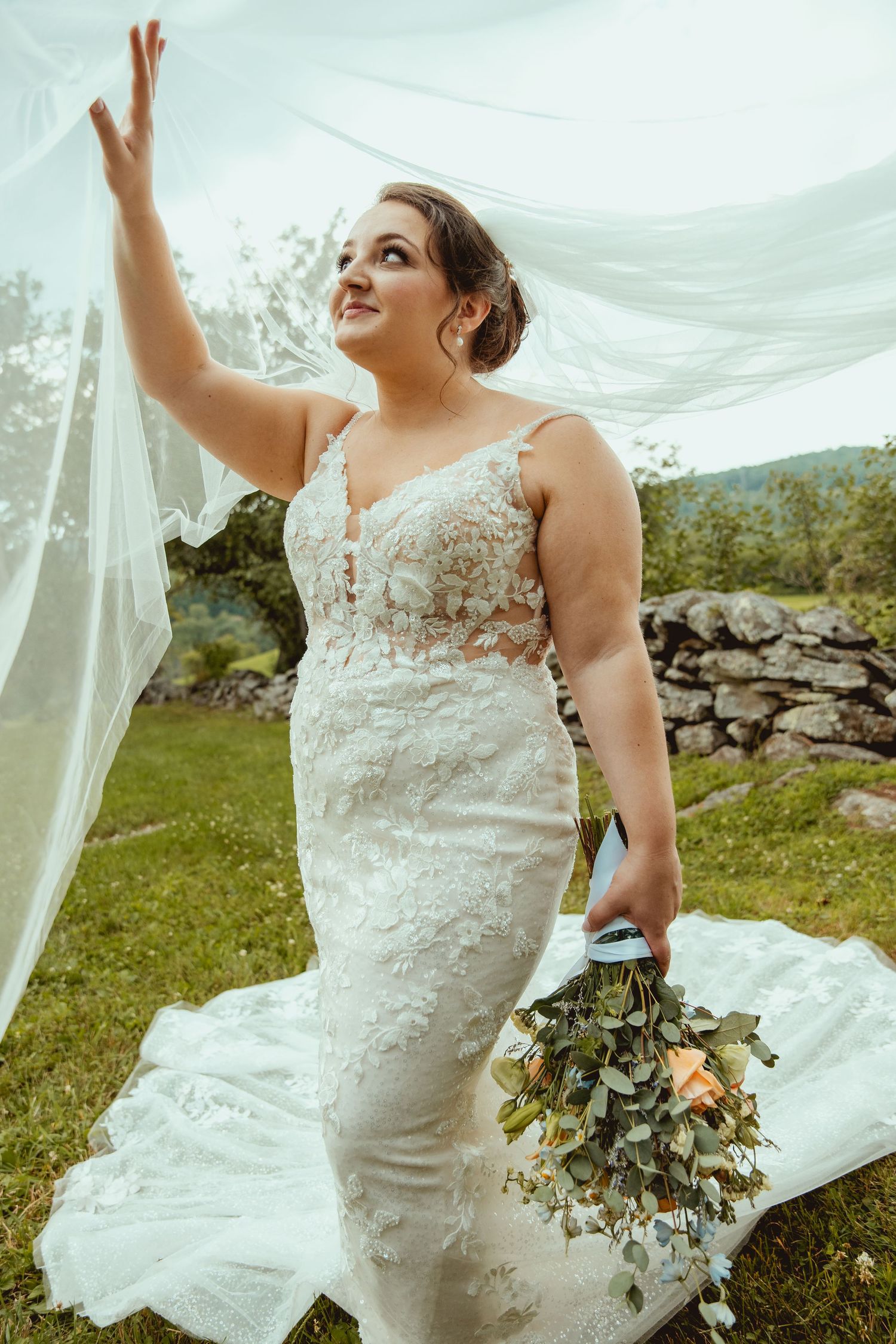 Bride in lace mermaid gown holds bouquet while veil flows overhead in outdoor garden setting.