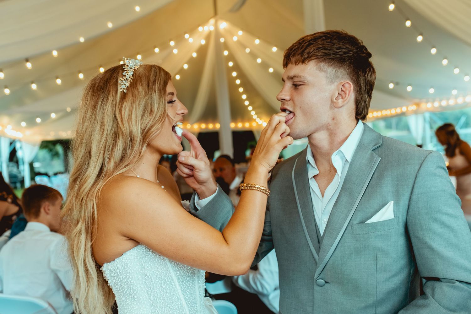 Newlyweds share wedding cake at elegant tent reception with string lights.