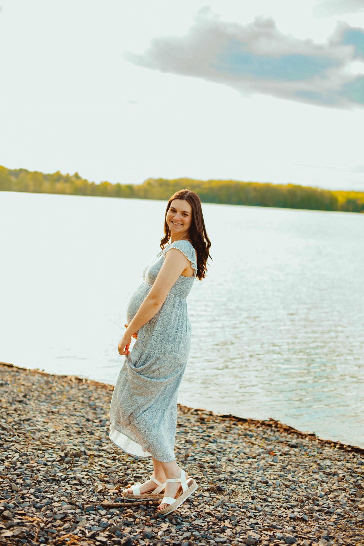Someone in a flowing light blue summer dress stands on a rocky lakeshore during golden hour.