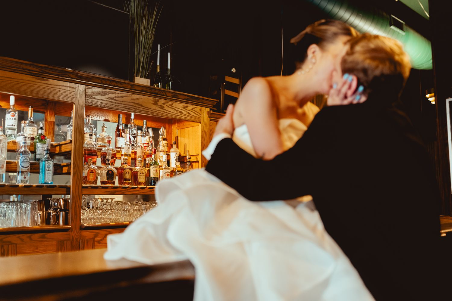 A bartender in a white shirt pours drinks at a dimly lit bar counter with bottles visible in the background.