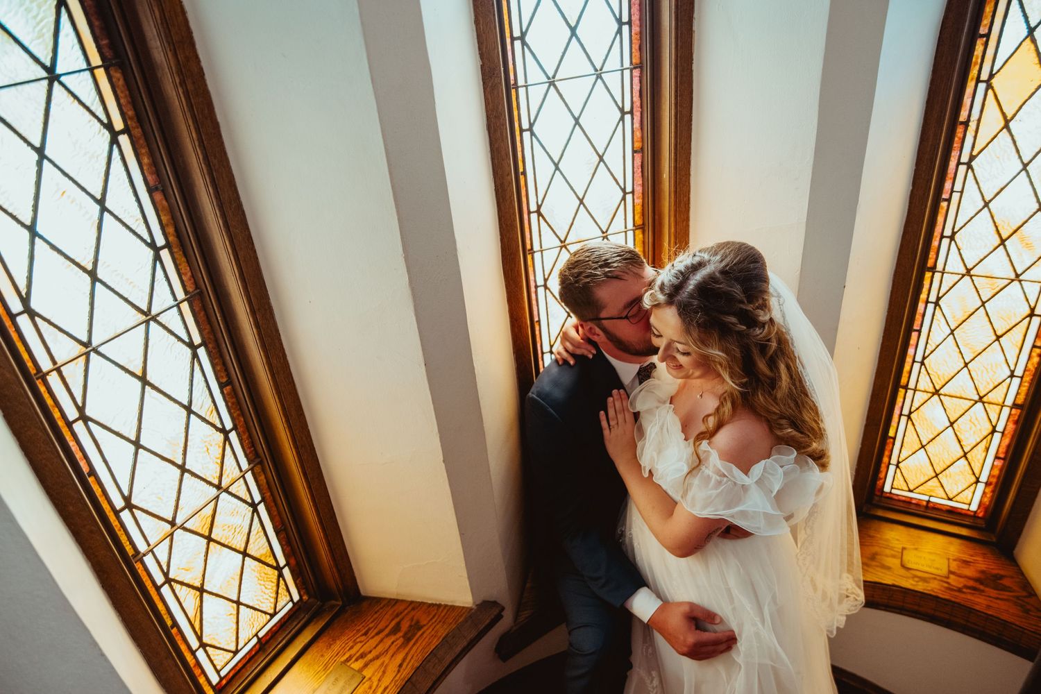 Bride and groom share a romantic embrace by ornate stained glass windows in a sunlit wedding venue.