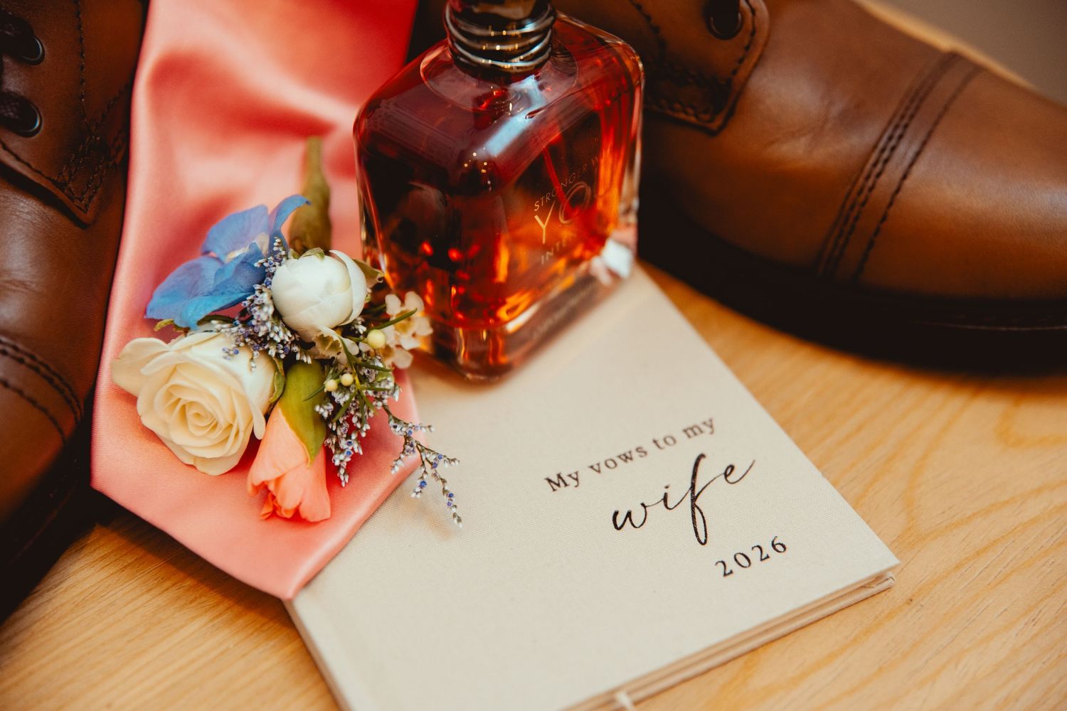 Wedding details flat lay featuring vows booklet, cologne, boutonniere, and brown leather dress shoes.