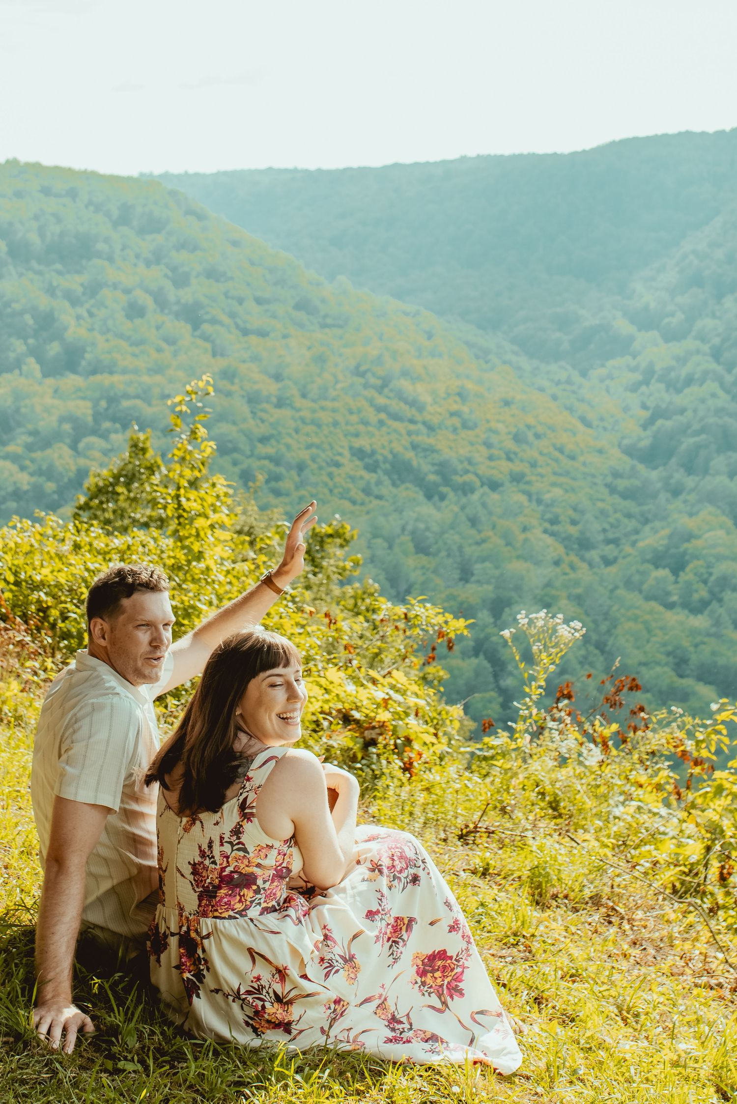Couple sitting on a scenic mountain overlook, woman in floral dress pointing at lush green valley views.